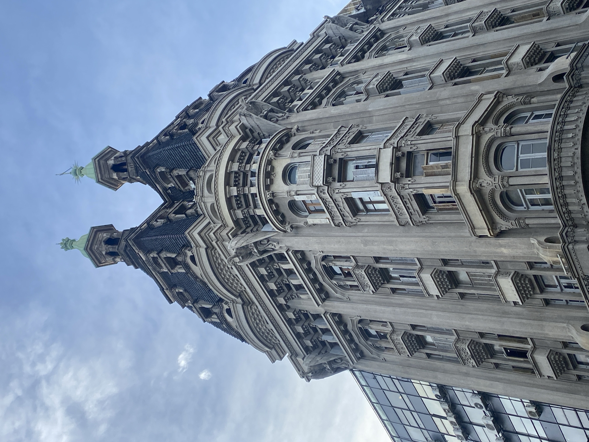 A low-angle shot presents the Palacio Barolo, located in Buenos Aires, Argentina. The architecture is a blend of Italian and Gothic styles, with twin towers capped by green spires, ornately decorated facades, and numerous windows. The sky is overcast with patches of blue, and a modern glass building is visible in the lower right corner.