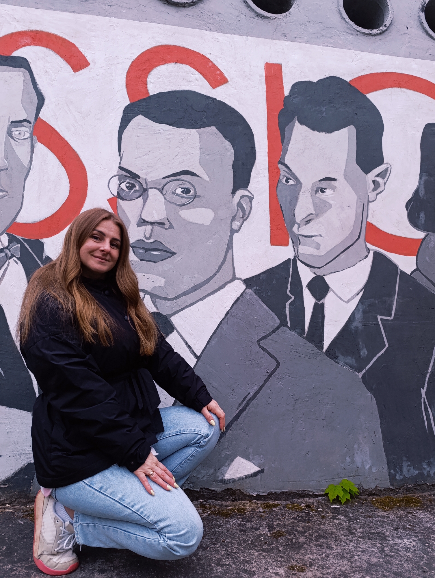 A young woman with long brown hair is crouching in front of a large mural depicting several men in suits. The mural features the letters "SSIO" in large red characters. The woman is wearing a black jacket and light blue jeans, with her hands resting on her knees. The background consists of a wall with the mural, and the foreground is a concrete surface with a small plant growing at the base of the wall. The location is the city of Unknown, Unknown.