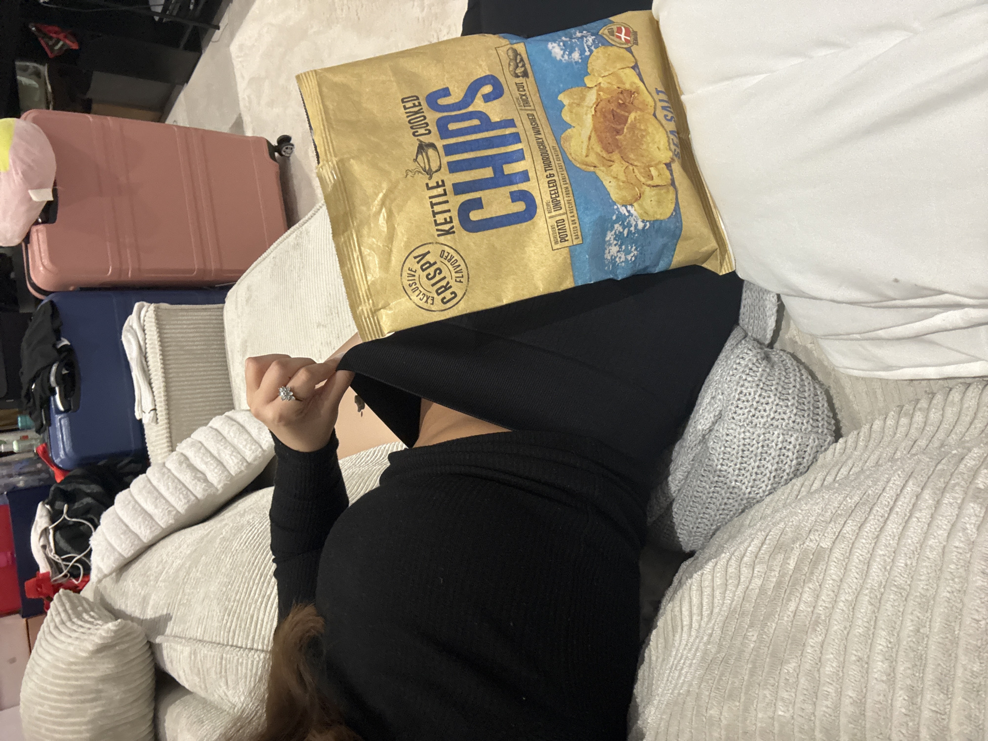 A person is lying on a light-colored corduroy couch, with a bag of Kettle Cooked Chips positioned over their face. The bag is yellow and blue, with an image of potato chips and text indicating "Sea Salt" flavor. The person is wearing a black long-sleeved top. Their left hand is visible, with a ring on the ring finger. Next to the couch is a white pillow and a grey knitted pillow. Behind the couch are luggage and other items. The location of the image is not discernible.