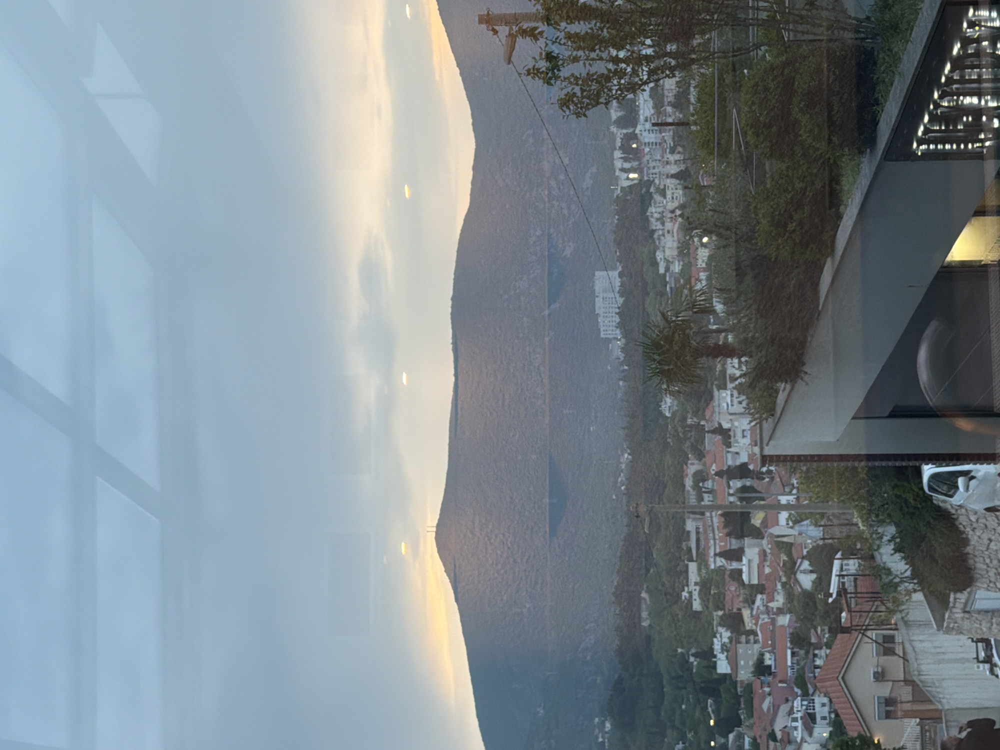 The image shows a view from a building in Mostar, Bosnia and Herzegovina. In the background is a mountain range. In the foreground, one can observe a residential area with buildings and trees. The lighting suggests it is either sunrise or sunset. There is a reflection of the building interior on the left side of the image. A construction crane is visible on the mountain side.