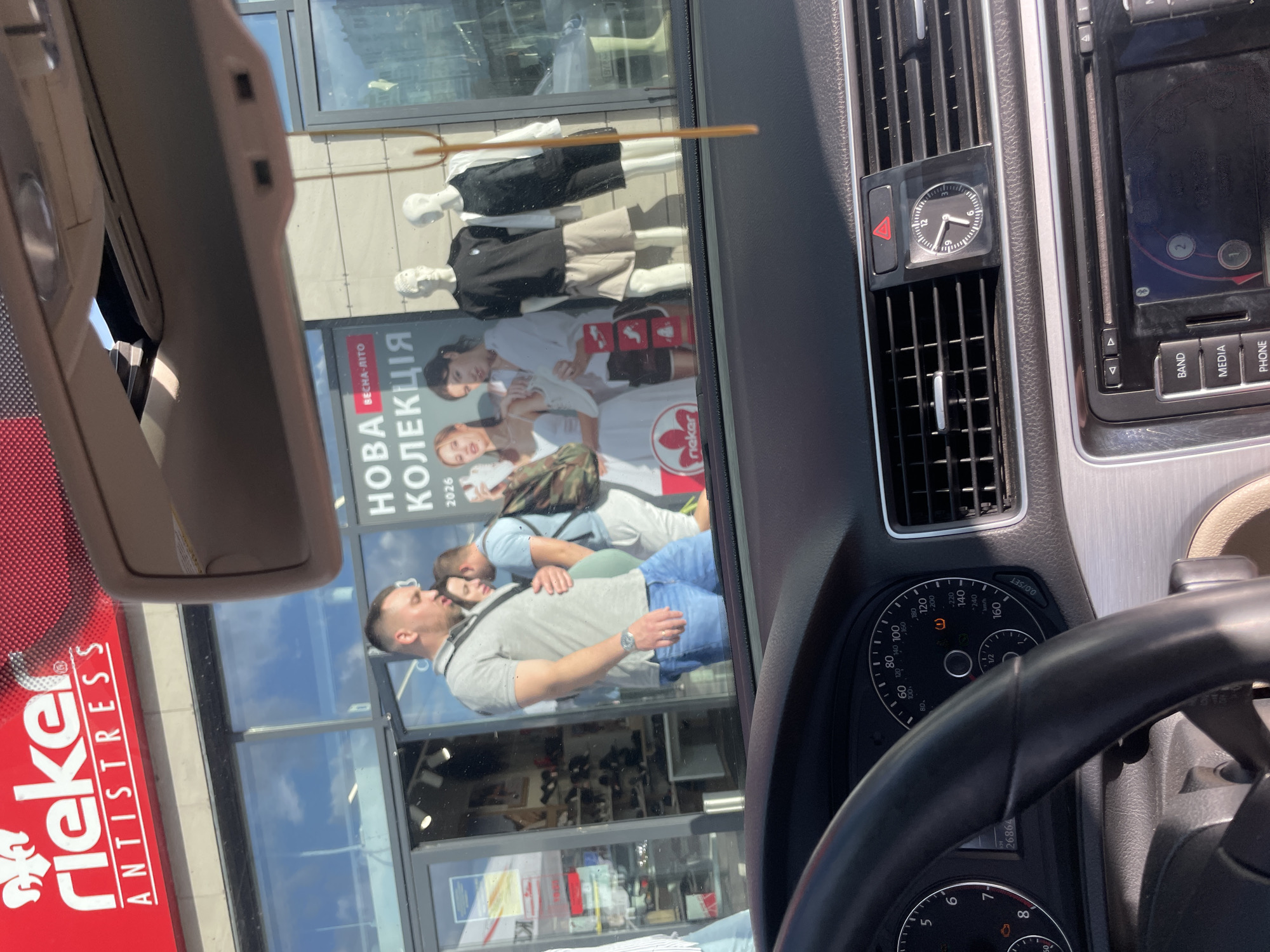 This media file captures a view from inside a car, looking out through the windshield. The scene is reflected on the glass, showing a street view with a storefront. The storefront features mannequins dressed in casual clothing, suggesting a clothing store. A large advertisement on the storefront prominently displays the text "НОВА ВЕСНА-ЛІТО КОЛЕКЦІЯ 2026" (New Spring-Summer Collection 2026) and the brand name "rieker" with "ANTISTRESS." There's also a reflection of people, possibly customers or passersby, interacting near the storefront. Inside the car, the dashboard is visible, including a clock on the air vent showing approximately 9:05, and part of the steering wheel and instrument cluster, indicating the car is likely parked or moving slowly. The lighting suggests it is daytime, possibly late morning or early afternoon, with bright sunlight. The overall impression is a glimpse of everyday life in a city, likely Kyiv, Ukraine, given the text in Ukrainian.