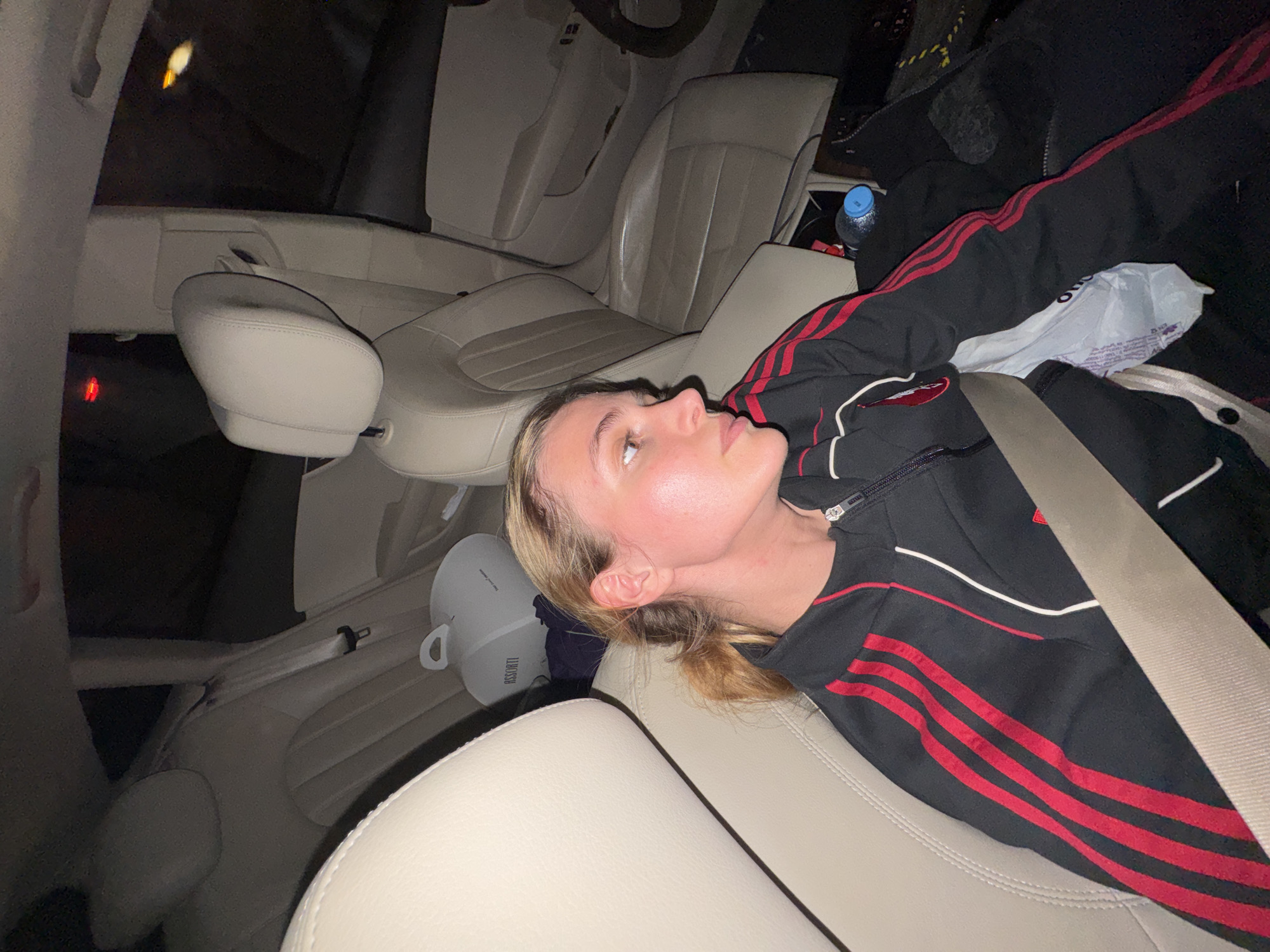 A young woman is depicted resting or lying down in the backseat of a car, facing upwards and towards the right of the frame. She has blonde hair pulled back, and her complexion appears fair with a slight flush on her cheeks. She is wearing a black tracksuit with prominent red stripes on the sleeves and body, and a white stripe detail on the chest, along with a silver zipper. A seatbelt diagonally crosses her torso, securing her.
The car's interior features cream or tan leather seats, which are visible in both the front and back. The car's overall interior seems dark, suggesting it is nighttime, with artificial lighting illuminating the cabin. The front passenger and driver seats are visible ahead, with a steering wheel partially seen in the distance on the left.
Various objects are present in the car: a blue-capped plastic water bottle rests on the console area between the front seats, and a white plastic bag is next to it on the rear seat. Another object, a white cylindrical container with a handle, possibly a humidifier or cooler, is visible behind her head on the seat, bearing the text "RASSORTI". A dark jacket or piece of clothing is also draped over the seat next to her. A faint red light is visible in the distance on the left side of the frame, and a yellow light is visible at the top. The scene takes place in Tbilisi, Georgia.
