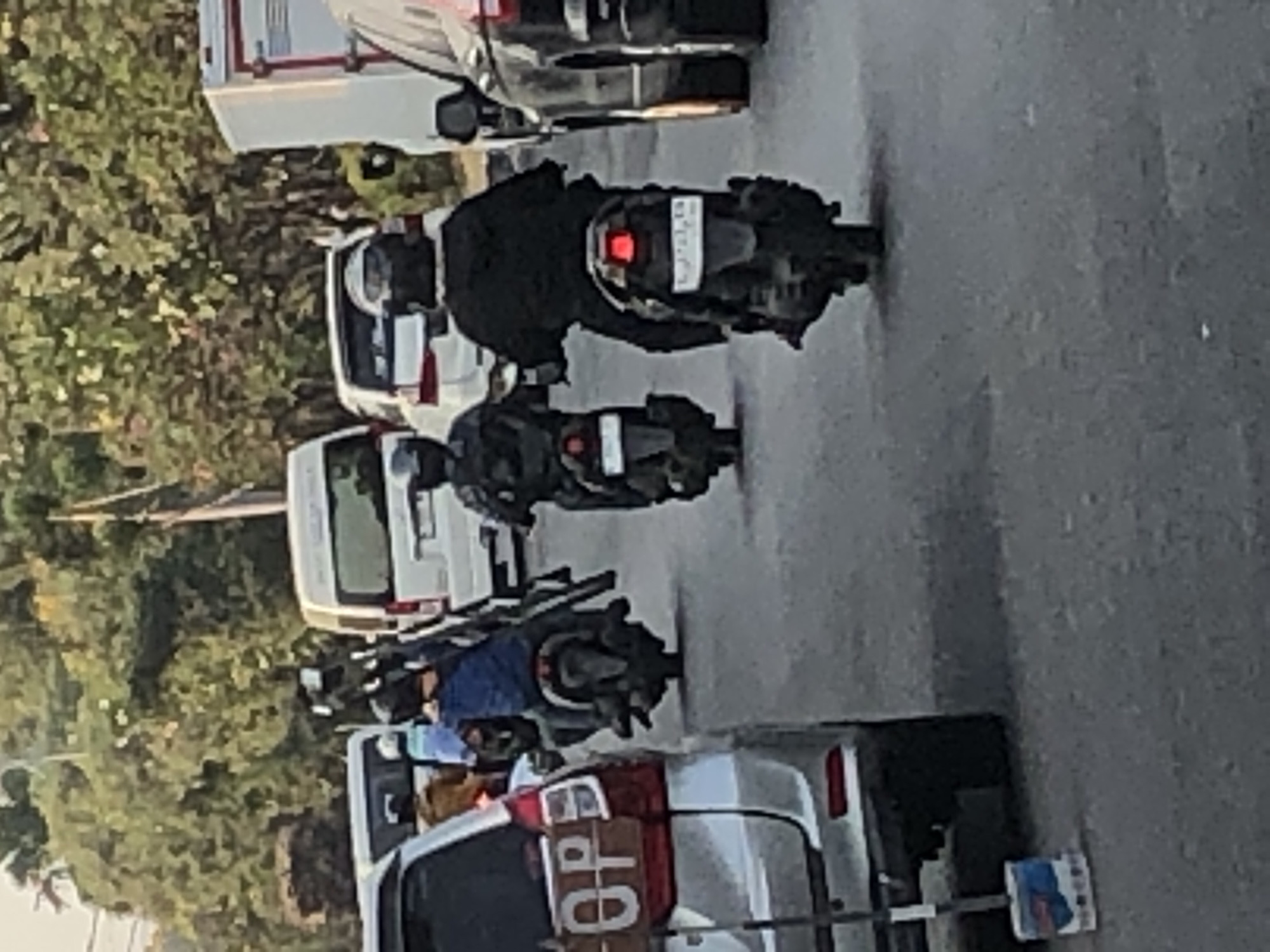The image shows a street scene in Ngaban, Indonesia, with several vehicles and people. A silver SUV is parked on the left side of the road, with a sign on its rear that says "OPP". Behind the SUV, two people on motorcycles are visible. The first person is wearing a blue shirt and riding a dark motorcycle. The second person, further down the road, is wearing a black jacket and riding a similar motorcycle. In the background, there are several white vans, a truck, and dense foliage on the side of the road. The sky is overcast, suggesting it might be late afternoon or early morning. The road surface appears to be asphalt, and there is some debris on the ground, including a mop on the left.