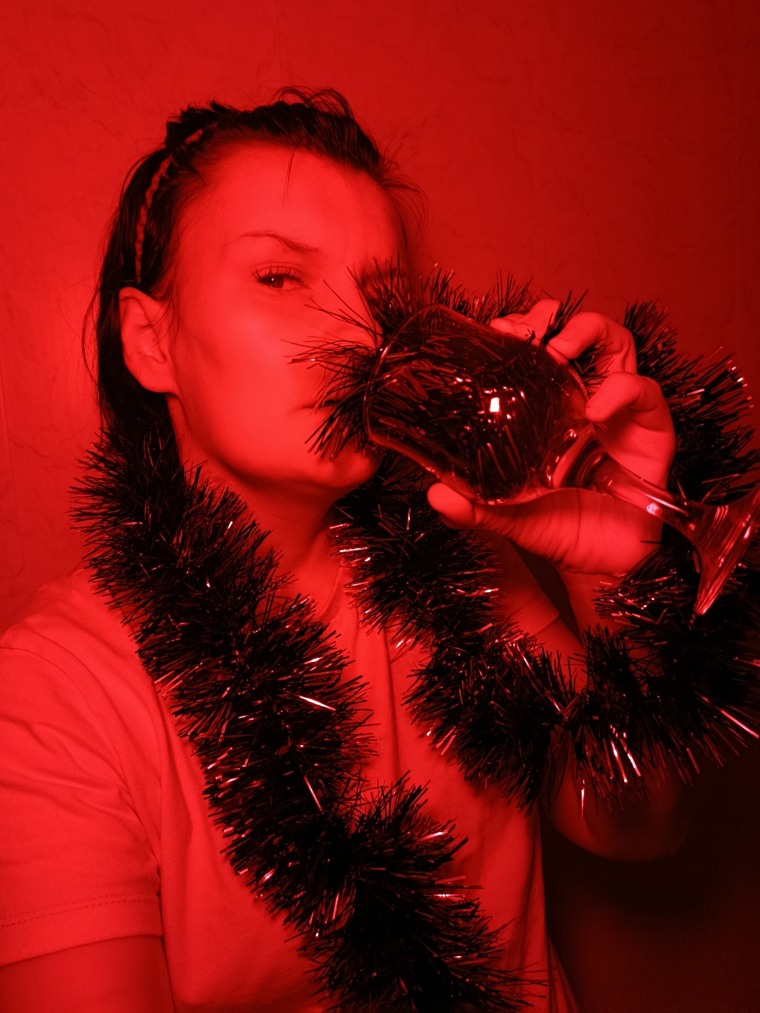 A woman is holding a wine glass to her lips, seemingly drinking from it. She is wearing a tinsel garland around her neck. The image has a strong red tint. The location is not discernible.