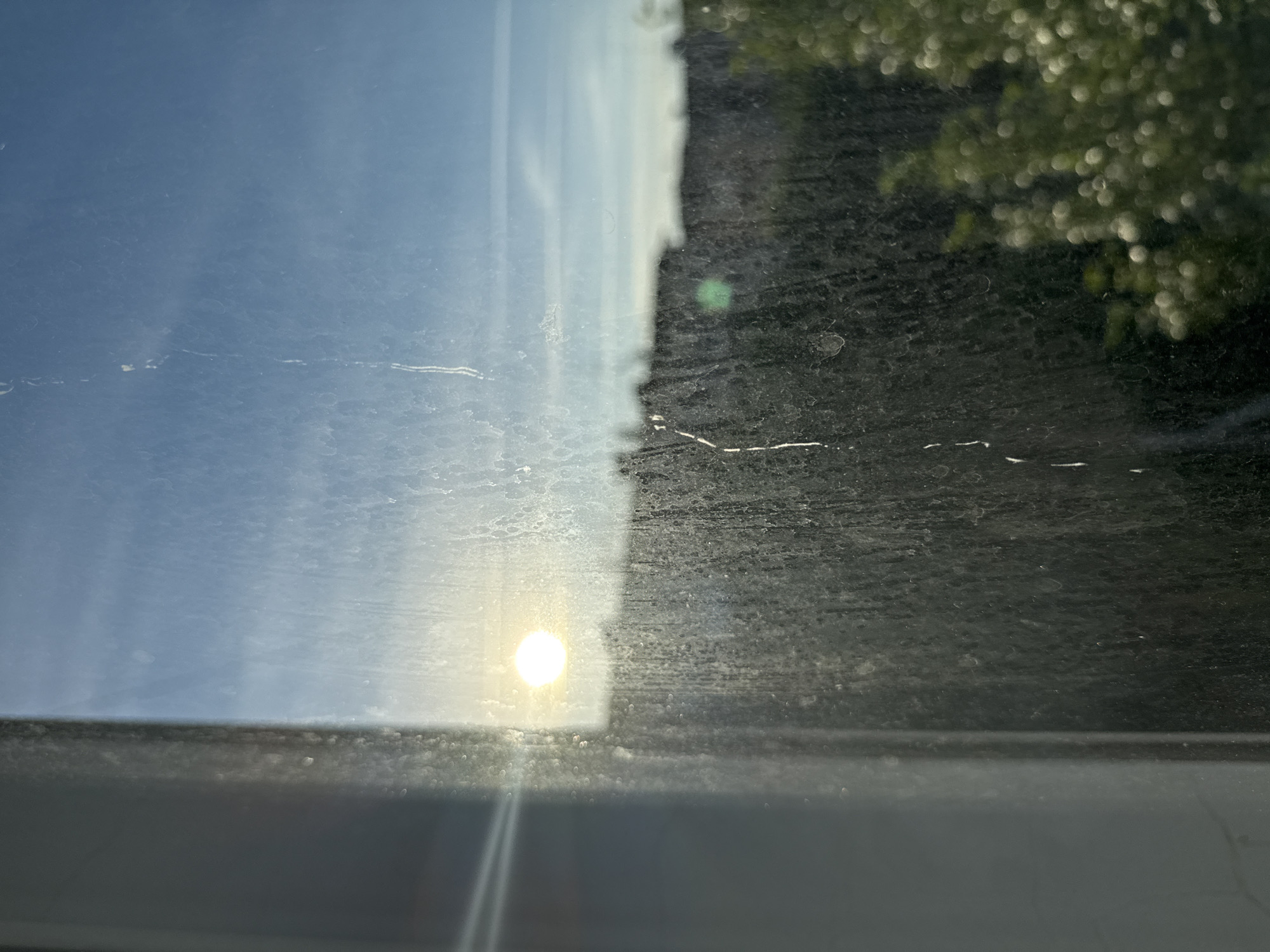 The image shows a view through a dirty window in Volgograd, Russia. The sun is visible in the sky, creating a bright glare. In the distance, buildings and trees can be seen. The window appears to be in a residential building, likely an apartment. The dirt on the window creates a blurry, distorted effect on the view outside. The overall impression is one of urban decay and neglect, with the natural beauty of the sky and trees being obscured by human-made grime.