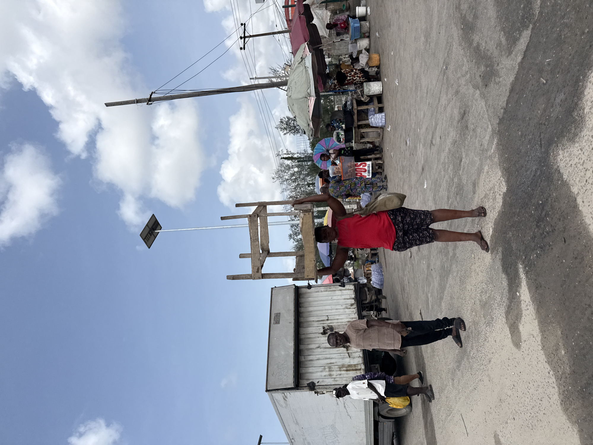 A daylight scene in Lagos, Nigeria. A woman in a red shirt and patterned shorts carries a wooden structure atop her head. Two men stand near a white truck. A market is visible in the background with vendors and customers. The sky is partly cloudy. A utility pole is visible. The surface is paved with rough cement.