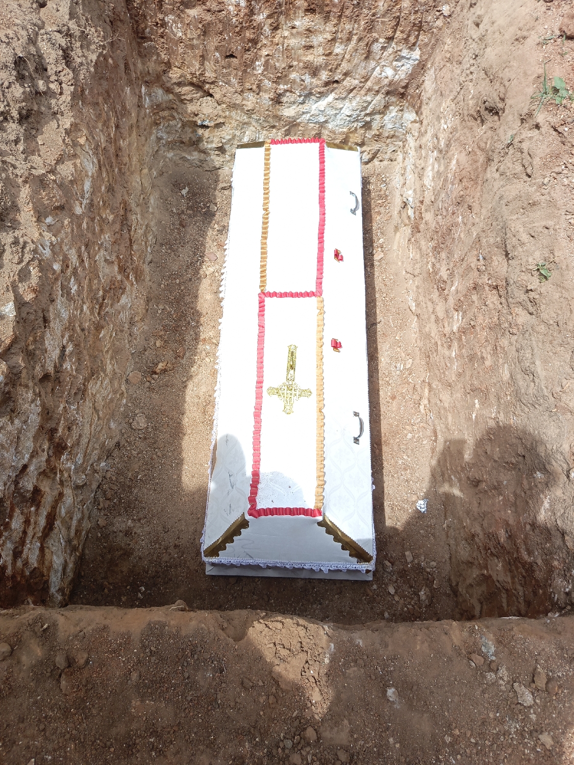 A white coffin is depicted lowered into a rectangular grave, viewed from above. The coffin is predominantly white, adorned with decorative gold and red trim along its edges and across the lid, forming a cross shape in the center. A golden, ornate Christian cross is prominently affixed to the top of the coffin. The grave is dug into reddish-brown earth, with visible layers and textures along its vertical walls, suggesting a mix of soil and some rocky elements. Loose soil lines the edges of the grave opening. The scene is outdoors under bright daylight, as indicated by strong illumination and a faint shadow cast on the right side of the grave, possibly from the person taking the photograph. The overall context suggests a burial or funeral ceremony. The reddish-brown soil is consistent with the geological characteristics of Andaha, Nigeria. No text is visible in the image.