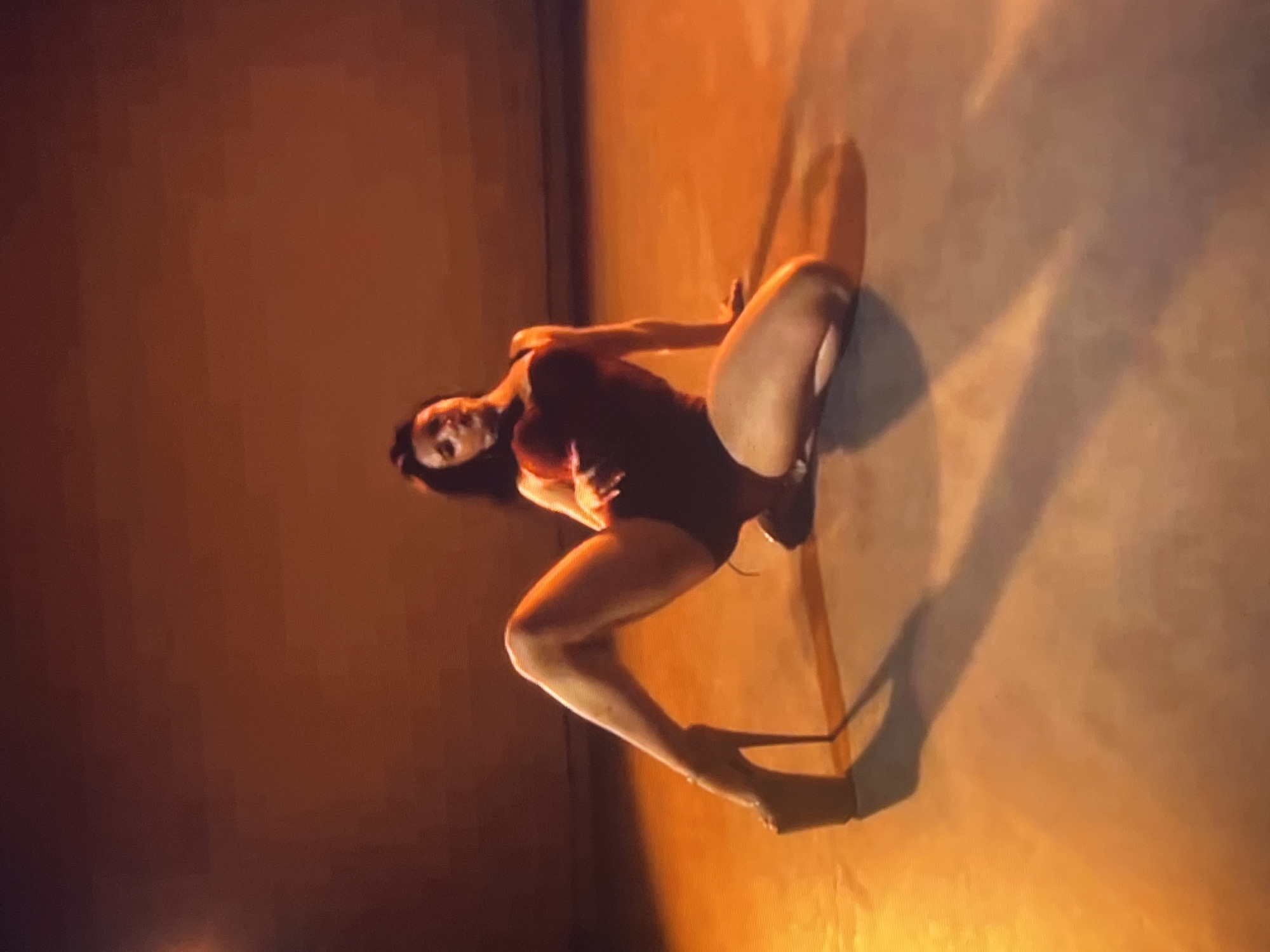 A woman wearing a dark red leotard and sky-high platform heels is posing on a concrete floor. Her body is contorted in a way that emphasizes her legs, one of which is extended and the other bent at the knee. The background is a muted orange, suggesting an indoor or nighttime setting. The lighting casts a long shadow of the woman onto the floor, highlighting her pose. There is no visible indication of the city of Zoriane, Ukraine, or any other specific location.