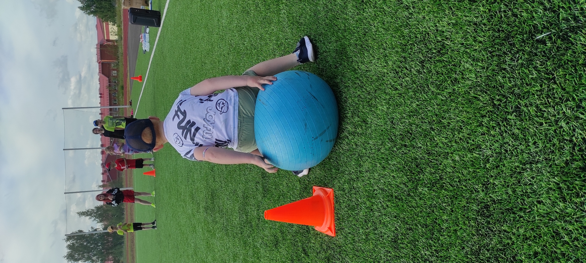 The image shows a boy wearing a black baseball cap and a white t-shirt with black graphics sitting on a large blue exercise ball on a green artificial turf field. He is facing away from the camera and his right hand is resting on the ball. He is wearing olive green shorts and black sneakers. To his left, there is an orange traffic cone. In the background, several children and adults are standing in a line, and a building with red roofs is visible behind them. The setting appears to be a sports or recreational facility in Noviy, Russia, likely a schoolyard or park.
