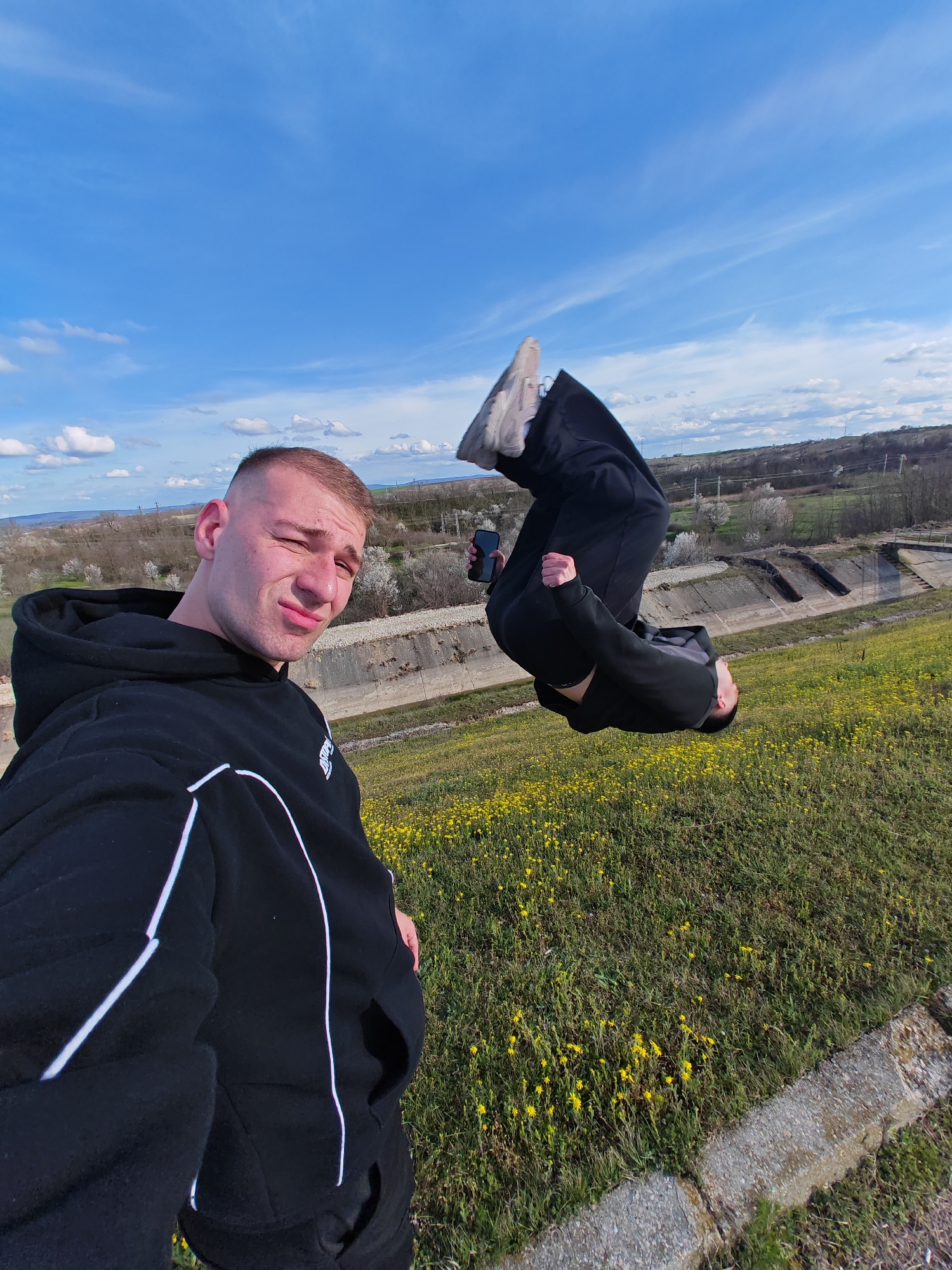 The image depicts a man taking a selfie while another man performs a backflip. The selfie-taker is on the left, wearing a black hoodie with white stripes and giving a slight smirk to the camera. The second man is mid-backflip on the right side of the frame, wearing a black outfit and light-colored sneakers; he is holding a black smartphone in his right hand. The background consists of a grassy slope covered with yellow flowers, a concrete structure resembling a drainage channel, and a landscape with scattered trees under a blue sky with scattered white clouds. The location is Shumen, Bulgaria.