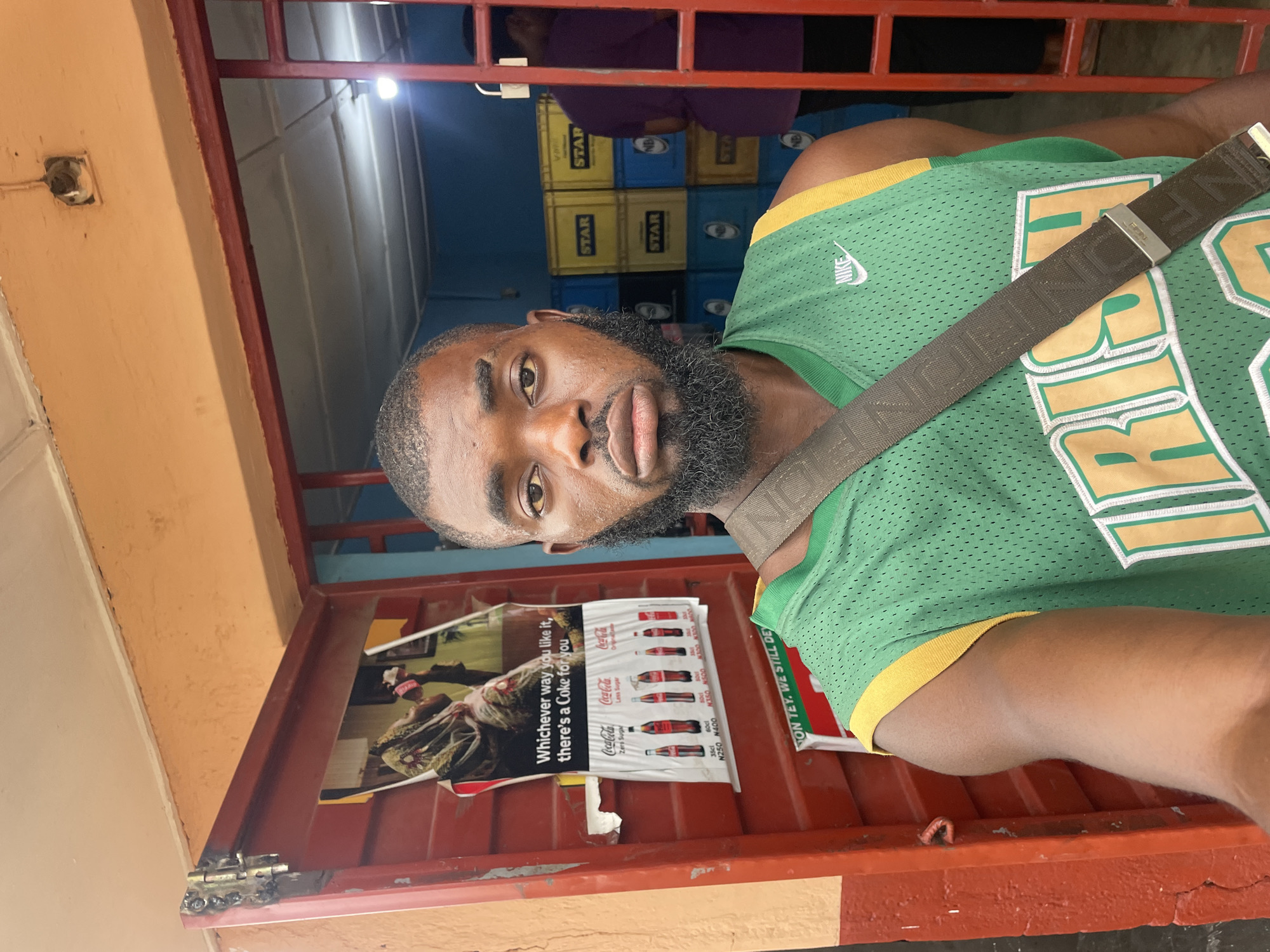 A young man with dark skin, a full beard, and short hair takes a selfie in front of a store. He is wearing a green basketball jersey with yellow trim and a Nike logo. A brown strap is visible across his chest. The store entrance has red metal bars and an orange-painted wall. A poster advertising Coca-Cola is visible on the wall inside the store. Behind the man, stacks of yellow and blue crates labeled "STAR" are visible. Another person in a purple shirt is partially visible in the background. The lighting suggests it is daytime. The scene appears to be in Jos, Nigeria.