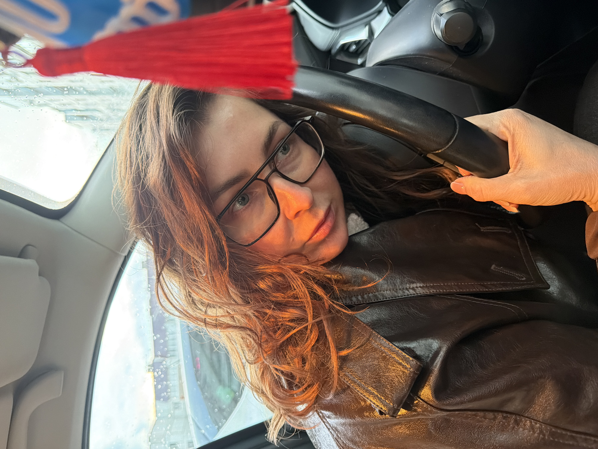 The image captures a selfie of a woman inside a car, seated in the driver's seat. She has shoulder-length, wavy, reddish-brown hair and wears black-framed glasses. Her right hand rests on the black steering wheel. She is dressed in a dark brown leather jacket. A vibrant red tassel is prominently visible hanging from the rearview mirror.
Through the car's windshield and side window, water droplets are evident, suggesting rain or wet conditions. The exterior view shows blurred outlines of buildings or structures, indicating an urban or suburban setting. There's also an unreadable blue and white logo or text visible through the top part of the windshield. The lighting on her face and hair is warm, possibly from sunlight breaking through clouds or an artificial light source. Her expression appears calm and neutral, as she looks directly at the camera. The setting is inside a car in Chaikovsky, Russia.