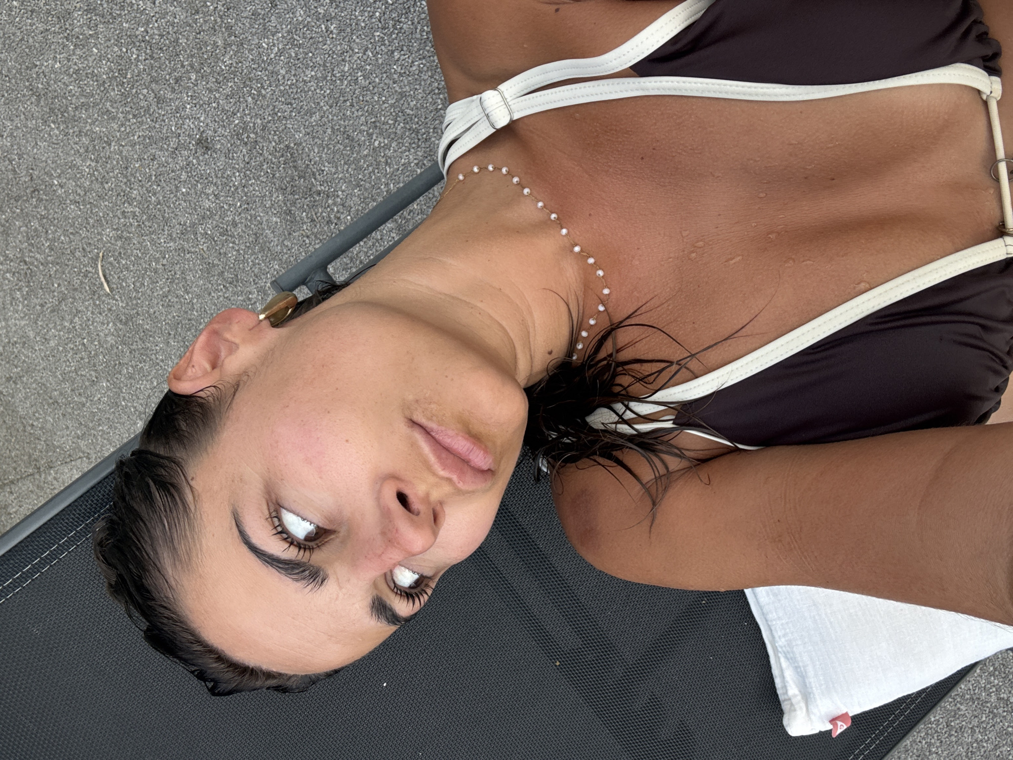 A woman is pictured lying on a recliner, facing upwards and to the left of the camera. She has dark hair, pulled back and wet, and appears to be outdoors. She is wearing a dark brown bikini with white trim and a delicate pearl necklace. Her skin is glistening with water droplets. The background consists of dark grey textured material, possibly a lounge chair, and grey gravel. The scene does not contain any elements that place it within the city of Veauchette, France.