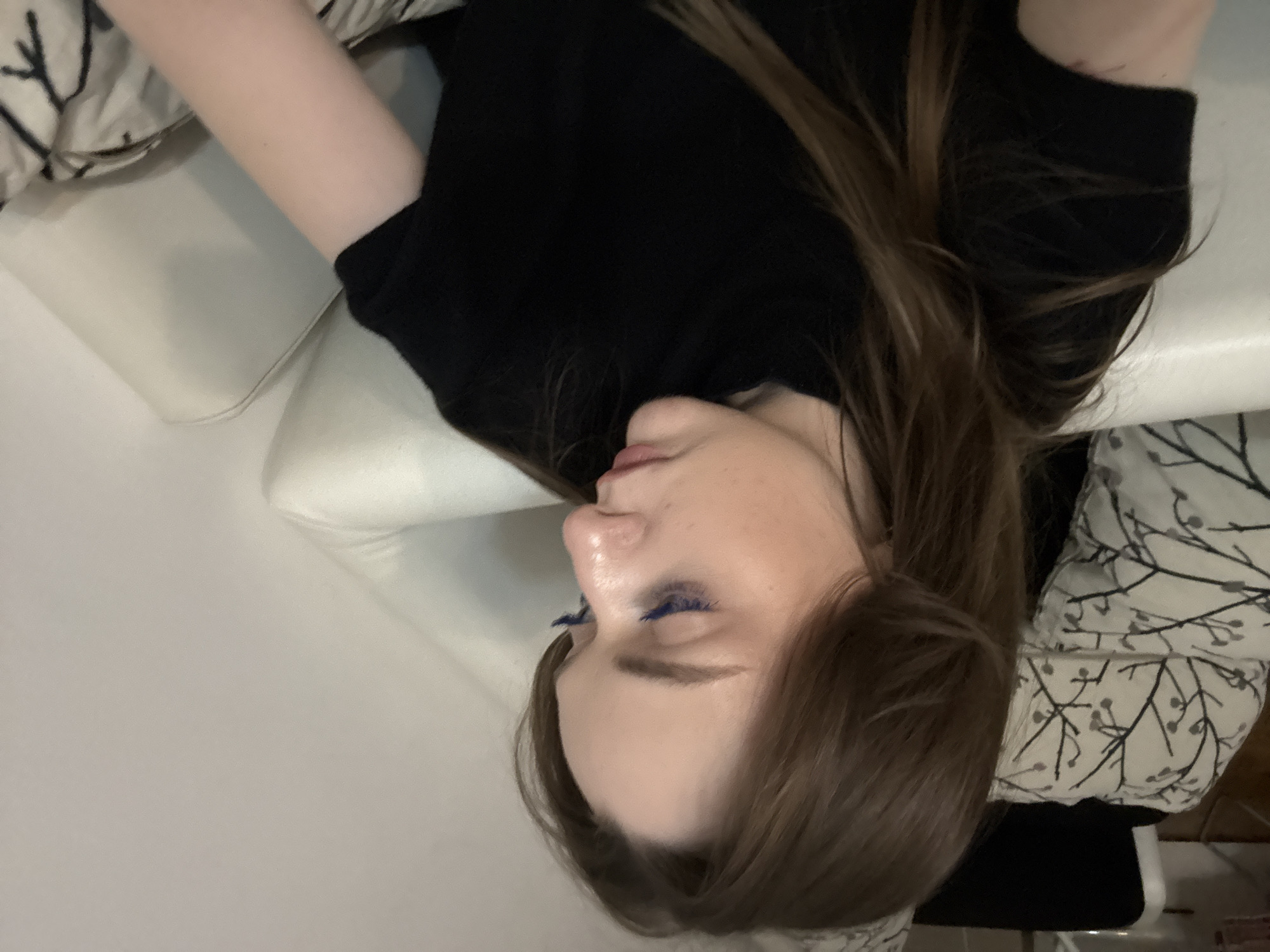 A woman with light skin is lying on a white leather sofa. She is wearing a black shirt. Her hair is brown and parted to the side. Her eyes are closed and have blue mascara. The background includes pillows with a black and white tree branch pattern. The location is unknown and cannot be determined from the provided image.