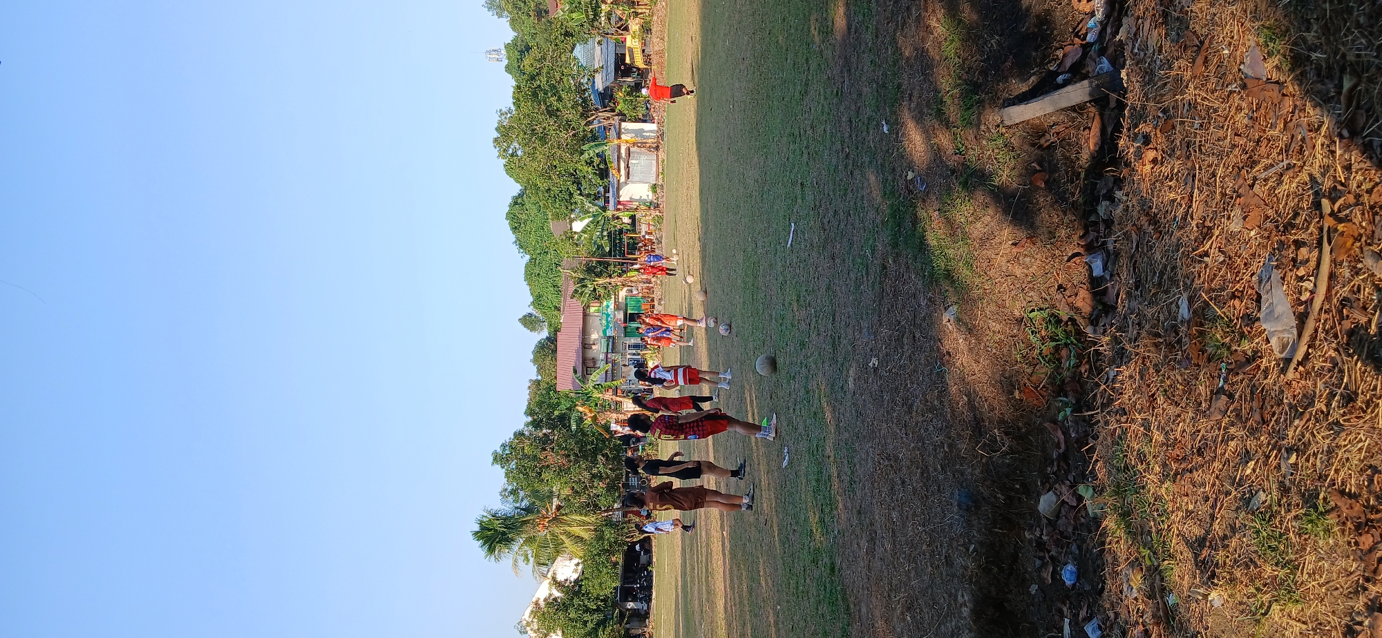 A group of people, likely a soccer team, are gathered on a dry grass field in front of buildings and trees. The sky is clear and blue. Several individuals are wearing athletic attire, with some standing near soccer balls. The ground is sparsely covered with grass and appears dry and somewhat unkempt, with some debris visible in the foreground. The scene appears to be a training session or casual game taking place in the afternoon. The background shows residential buildings and lush green trees, indicating an urban or semi-urban environment.