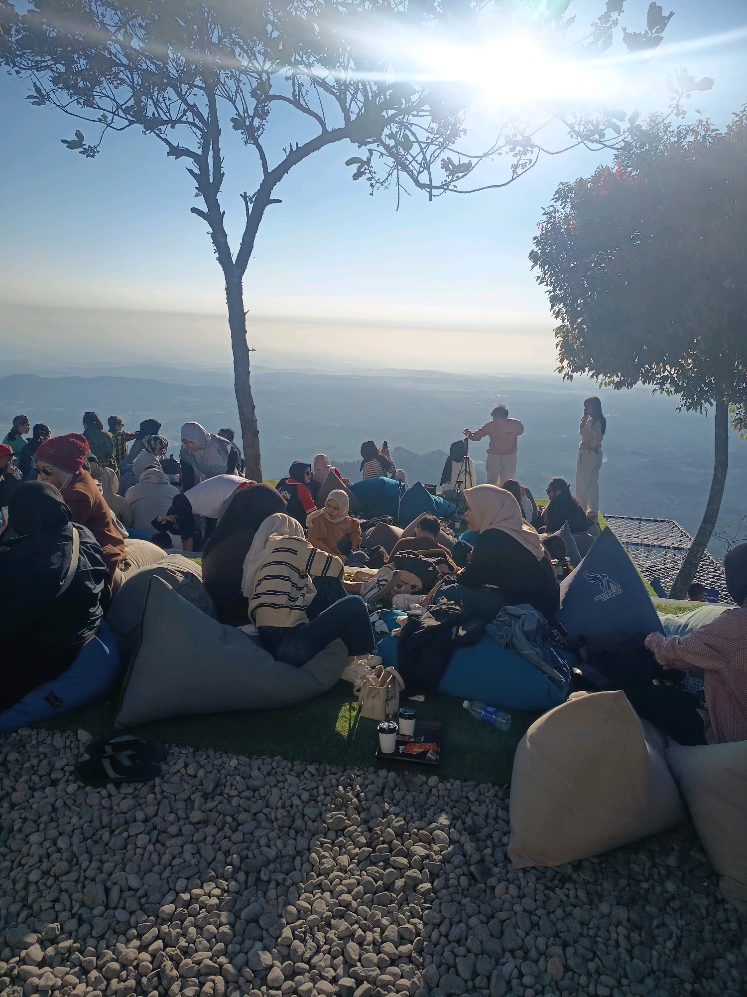 This image captures a group of people gathered outdoors on a grassy area overlooking a vast, hazy landscape, likely Keditan, Indonesia. The scene is bathed in bright, hazy sunlight, creating a lens flare effect. Many individuals are seated on beanbag chairs of various colors and sizes, spread across the foreground and middle ground. Some are also standing, including two individuals in the background who appear to be posing or interacting, one with a tripod setup nearby, suggesting photography or videography. The people are casually dressed, with many wearing headscarves. In the foreground, a section of the ground is covered in small, light-colored pebbles. Two trees frame the scene on the left and right, with one on the left being a bare, silhouetted tree against the bright sky and the one on the right featuring dense foliage. The distant background reveals rolling hills or mountains shrouded in atmospheric haze. The overall impression is one of a relaxed outdoor gathering at a scenic overlook.