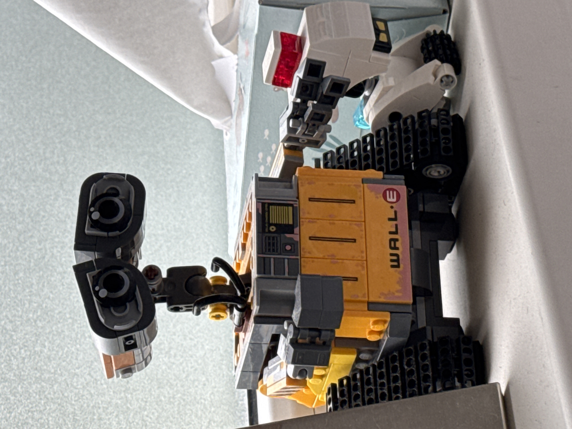 The image shows a close-up, eye-level, medium shot of a LEGO model of the robot WALL-E. The model is positioned on a white, textured surface, possibly a windowsill or shelf, with a blurred background that includes a box of tissues. WALL-E is depicted in its classic yellow and grey color scheme, with its distinctive binocular-like eyes and tank treads. To the right of WALL-E, a white LEGO creation with a red top light is partially visible, appearing to interact with WALL-E's arm. The lighting suggests it is daytime, with light coming from the left. There are no people or animals present in the image. The scene is indoors. The image does not provide any information or visual cues to suggest a location in Riga, Latvia.