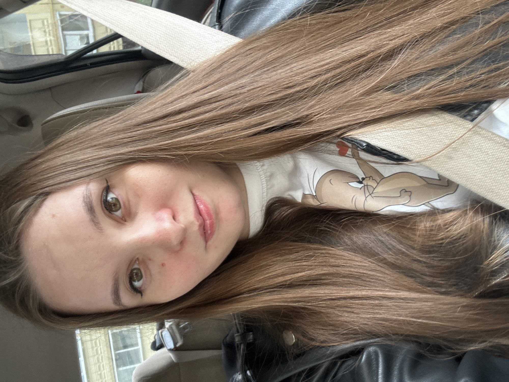 The image shows a close-up, vertically oriented selfie of a young woman in a car. Her face is turned towards the camera, with her eyes looking directly at it. She has long, straight, light brown hair that drapes across the frame, obscuring parts of the background. She is wearing a white t-shirt with a cartoon character printed on it. Her makeup includes eyeliner and lipstick. The background shows parts of the car interior, including a seatbelt, a black leather seat, and a glimpse of buildings outside the window. The lighting suggests it is daytime, and the scene appears to be taking place in Noviy, Russia. There is no visible text in the image.