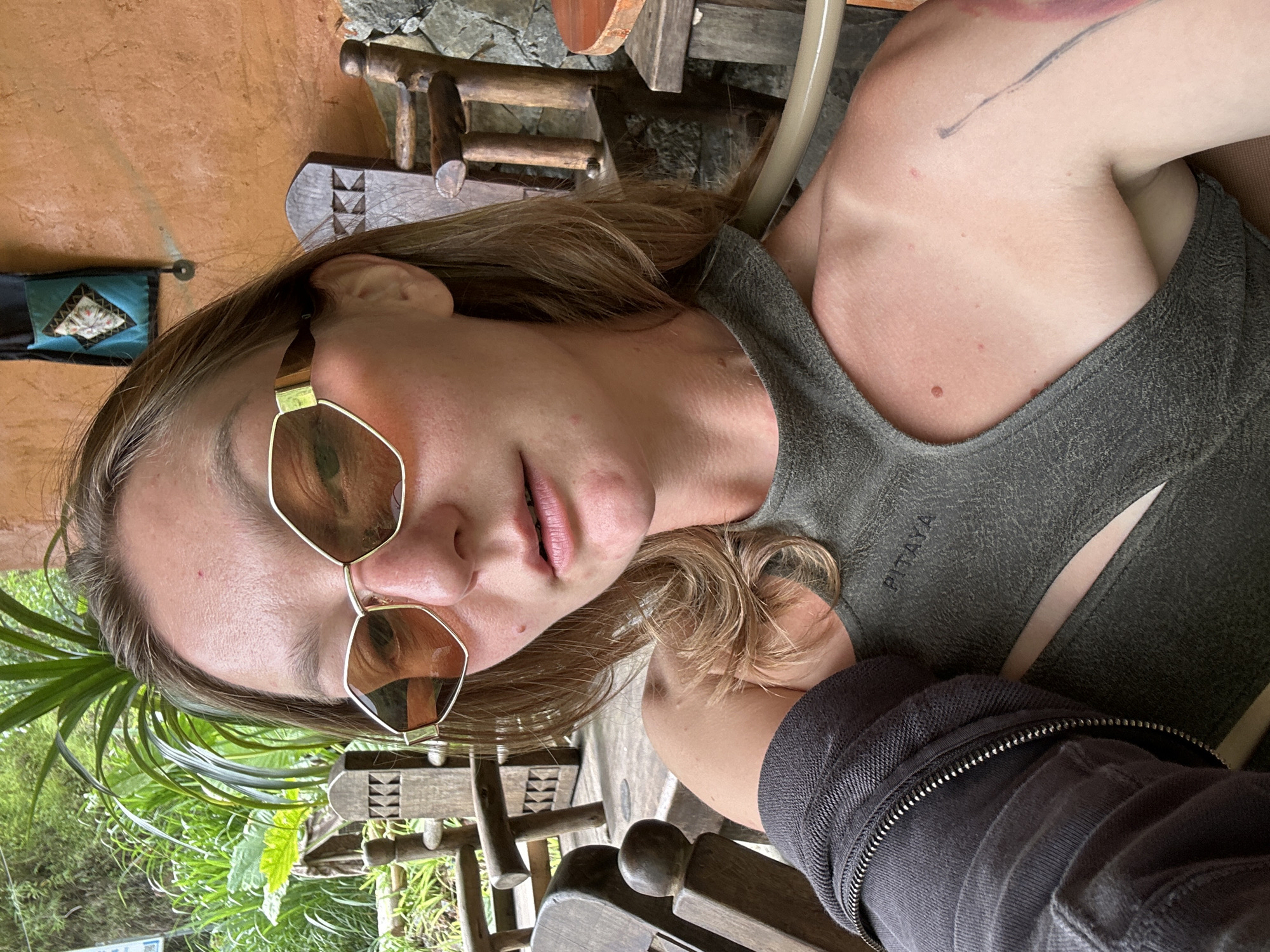 A woman is sitting on a wooden chair, wearing sunglasses and a green tank top with "PITAYA" printed on it, and a dark grey jacket. She appears to be outdoors, judging by the greenery visible in the background and the weathered wood of the furniture. The location appears to be in Yên Minh Commune, Vietnam, based on environmental clues. The scene seems peaceful and relaxed.