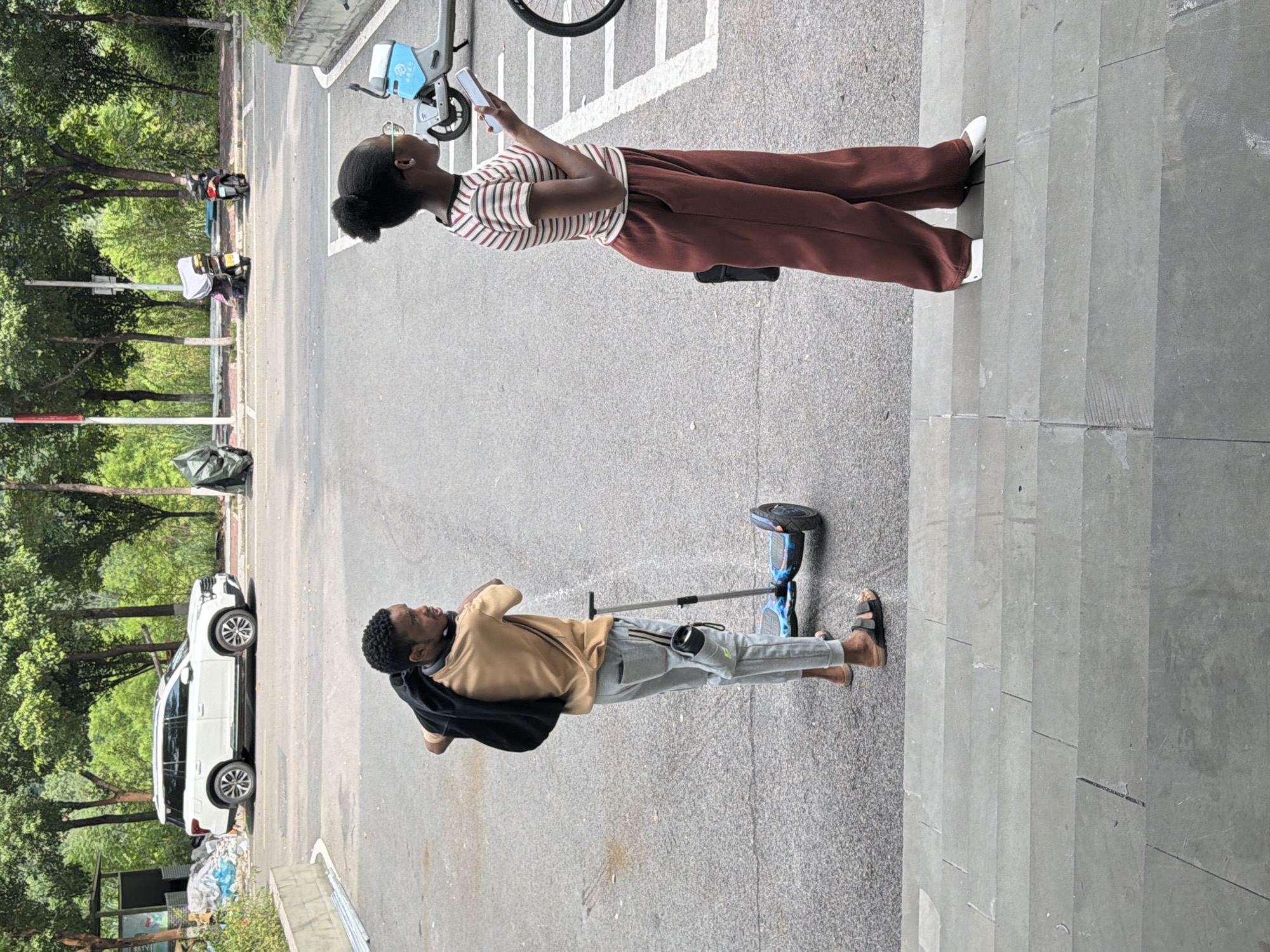 In Jiangjin, China, two individuals are in a paved outdoor area, possibly a parking lot or plaza, adjacent to a street lined with trees. On the left, a young Black man stands on a hoverboard, facing away from the camera. He is wearing a tan t-shirt, light gray sweatpants, and dark sandals. A black jacket is slung over his left shoulder, and a water bottle in a grey holder is attached to his right pant leg. On the right, a young Black woman stands, facing left, looking at her phone. She is wearing a horizontally striped t-shirt and loose-fitting, dark reddish-brown pants. She is also wearing white slip-on shoes. Behind the woman, a white electric scooter is parked. In the background, a white SUV is parked near a small, open-air structure. Further back, several parked scooters and more trees are visible. The ground is paved with asphalt, and large stone steps are in the foreground.