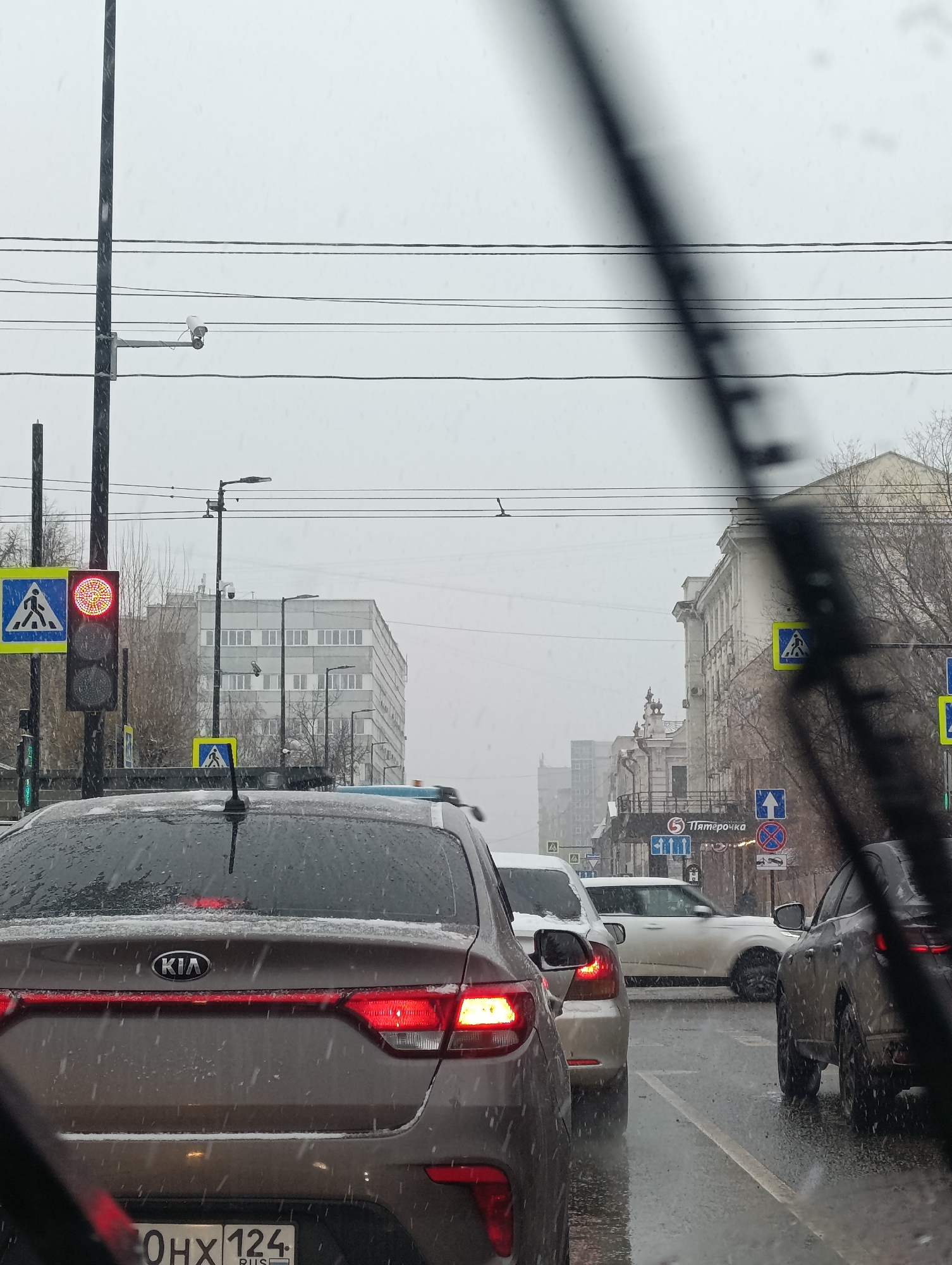 The image depicts a view from inside a car, looking out onto a city street in Krasnoyarsk, Russia, during snowy weather. Several cars are stopped at a traffic light with a pedestrian crossing signal showing red. A pedestrian crossing sign is visible beside the traffic light. The street appears wet. A surveillance camera is visible on a pole. Buildings line the street and traffic signs are clearly visible including a sign for the Pyaterochka supermarket. The license plate on the car in front is partially visible, indicating the region 124. The windshield wipers are partially visible.