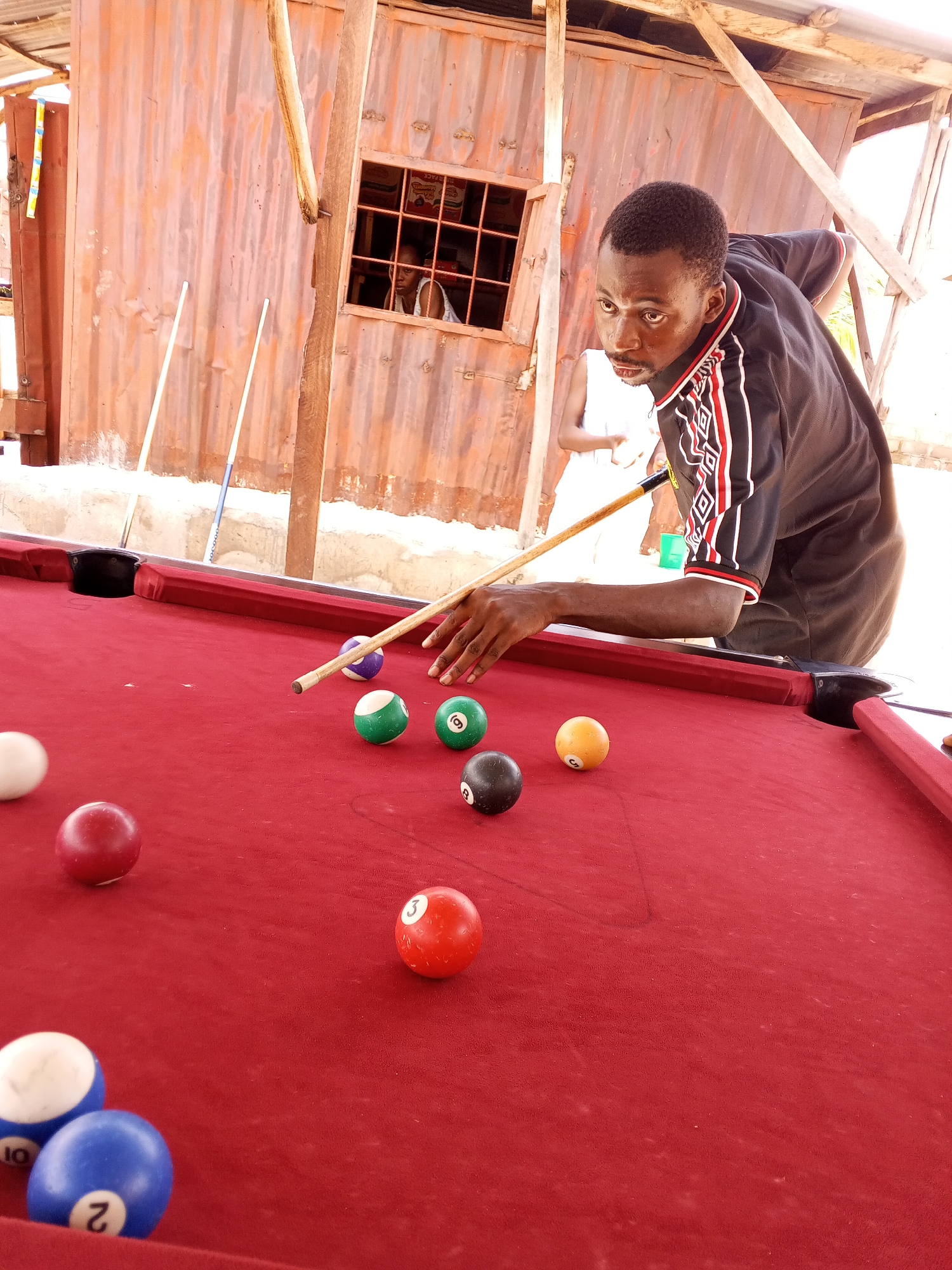 An adult male of African descent, wearing a black shirt with red and white trim, is playing pool on a red felt table. He is holding a cue stick and is in the process of striking a ball. Several pool balls are scattered across the table, including solid colors and striped balls. The game appears to be taking place outdoors, possibly in a makeshift establishment or a backyard, given the corrugated metal structure in the background and the presence of wooden poles. The background also shows two white poles leaning against the structure and a glimpse of another person through a window. The overall setting suggests a casual, informal environment. Based on the provided information, this scene is located in Lokoja, Nigeria.