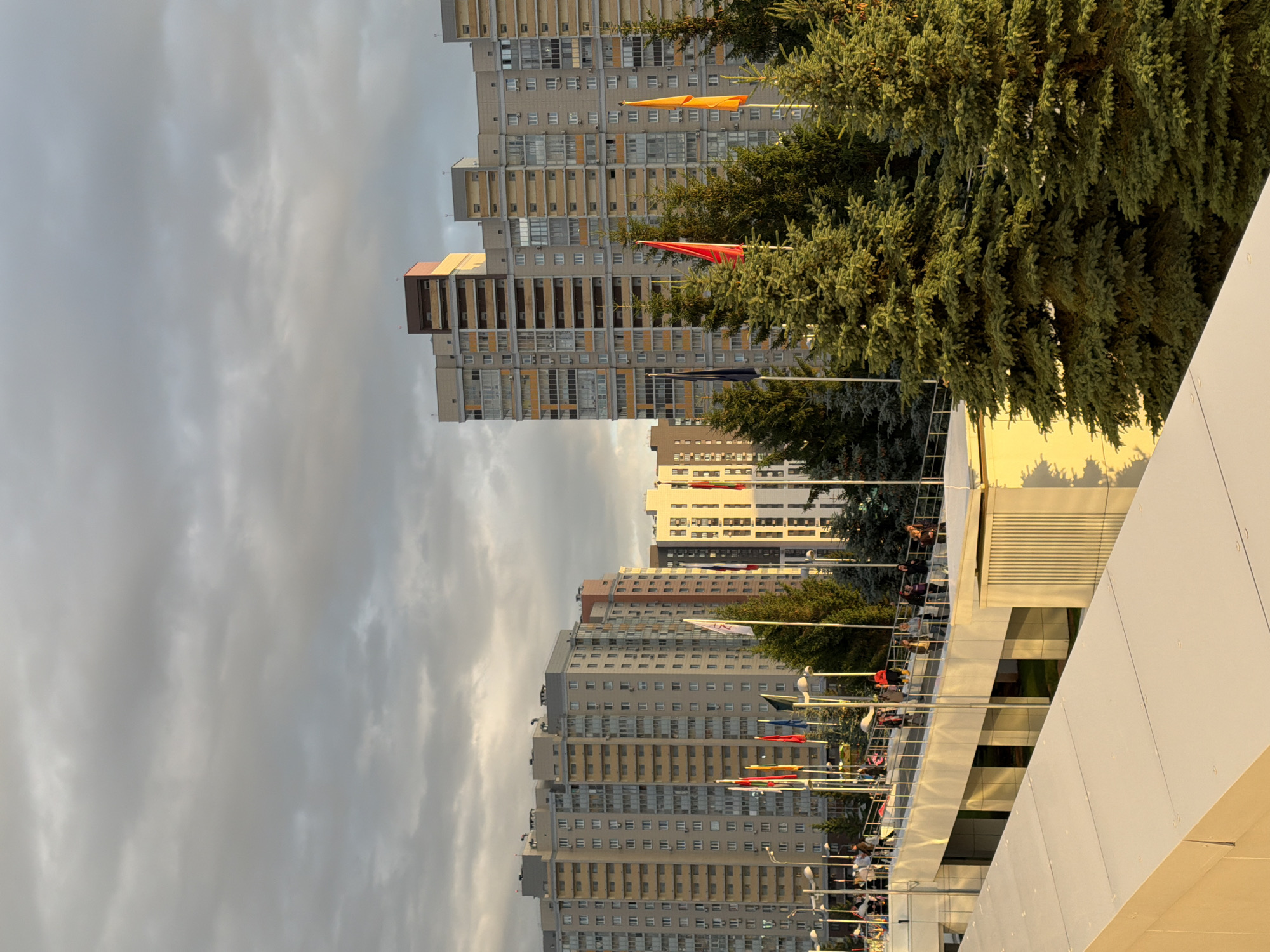 The image shows a city scene under a cloudy sky. In the foreground, there are two modern buildings, one with a light-colored angled roof and another with a cream-colored ribbed facade, both bathed in golden sunlight. A white pedestrian bridge with a metal railing extends across the midground, with several people walking on it. Behind the bridge, there are several tall, multi-story apartment buildings with light-colored facades and rows of windows. In front of the apartment buildings, a line of evergreen trees stands tall, interspersed with several flagpoles displaying colorful flags, including red, yellow, and dark blue. The overall lighting suggests it is either early morning or late afternoon.