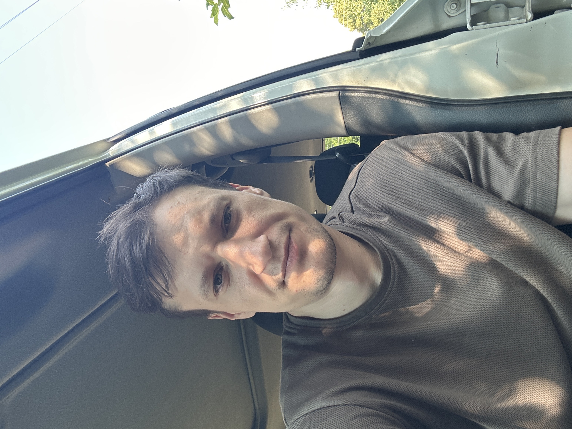 A man is sitting in the driver's seat of a car, looking at the camera. The car door is open, and sunlight is shining through the trees, casting shadows on the man and the interior of the car. The man has dark hair and is wearing a brown t-shirt. The scene appears to be in an outdoor setting, possibly a rural area or a park, given the presence of trees and sunlight. The location is Shatava, Ukraine.