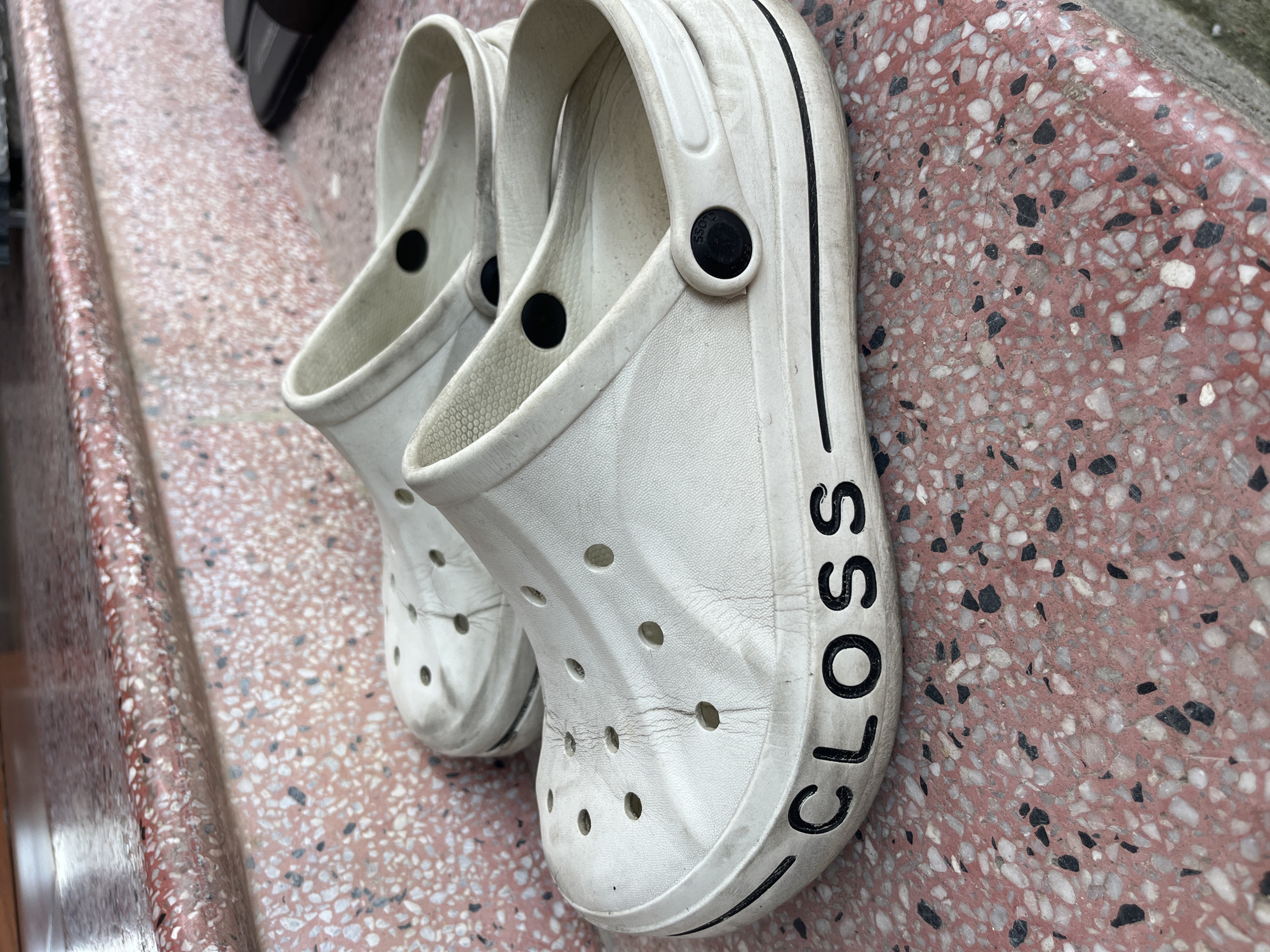 Two white "Crocs" brand clogs are placed on a speckled pink and grey surface. "Crocs" is stylized as "Closs" on the right clog. The surface appears to be part of an outdoor structure, possibly a step or ledge. This photograph was taken in Bac Nha Trang Ward, Vietnam.