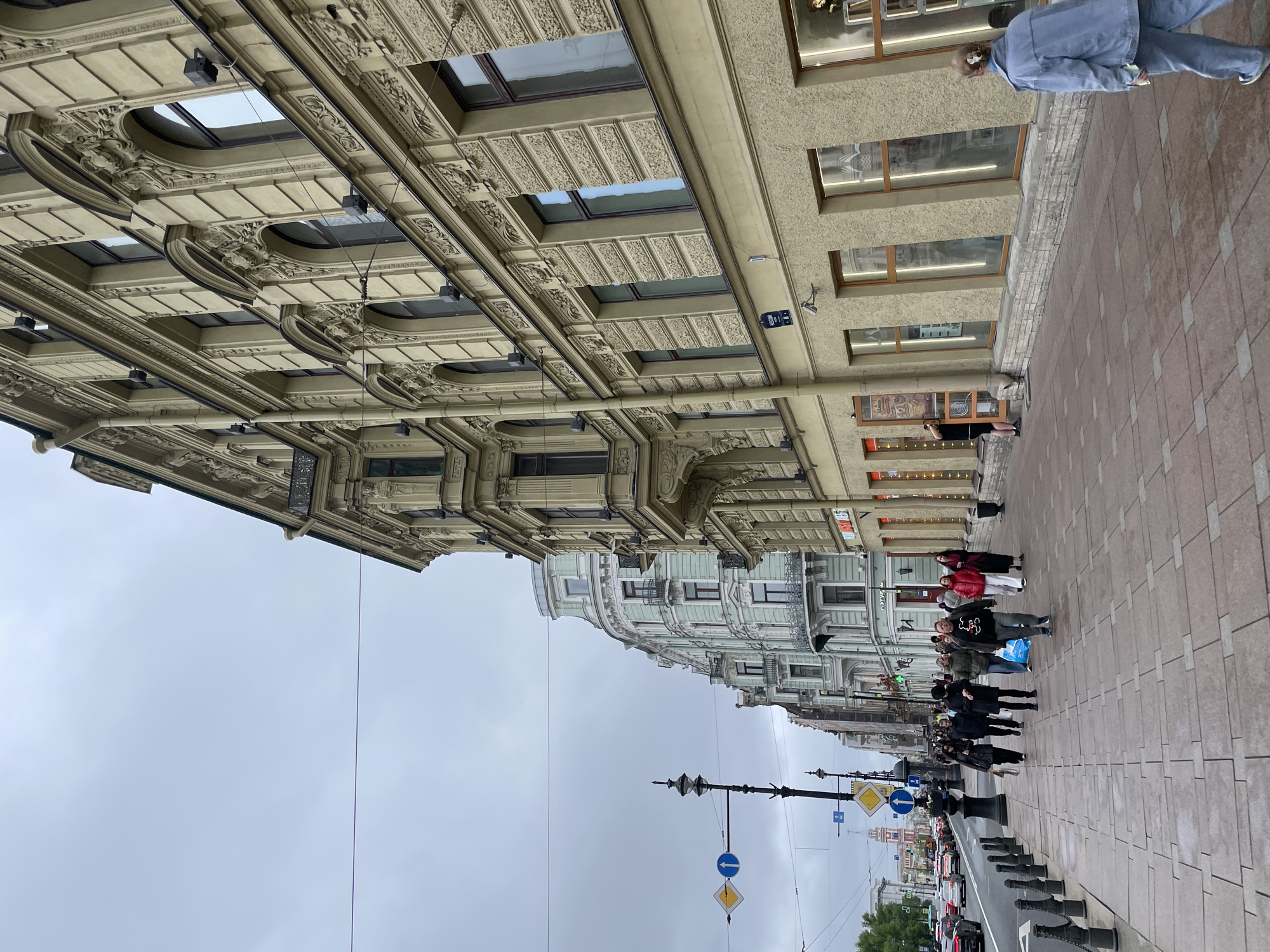 The image captures a street scene in Saint Petersburg, Russia, likely along a main thoroughfare. The camera is positioned at street level, looking along a pedestrian walkway adjacent to a busy street with automobile traffic. On the right side of the walkway is a row of commercial buildings with window displays. Pedestrians are walking along the sidewalk. The buildings on the left side of the walkway are older and more ornate with decorative facades and large windows. Overhead streetlights and traffic signs are visible. The sky is overcast, indicating a cloudy day.