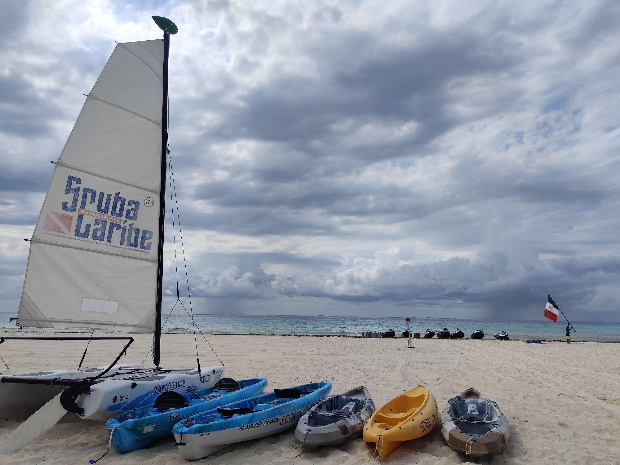 The image shows a beach scene in Playa del Carmen, Mexico, under a cloudy sky. In the foreground, a white and blue catamaran with the "Scuba Caribe" logo on its sail sits beside four kayaks of various colors (blue, gray, and yellow). Further back, on the sand near the ocean, are several jet skis lined up. In the distance, a person stands holding a flag with the colors of the State of Mexico.