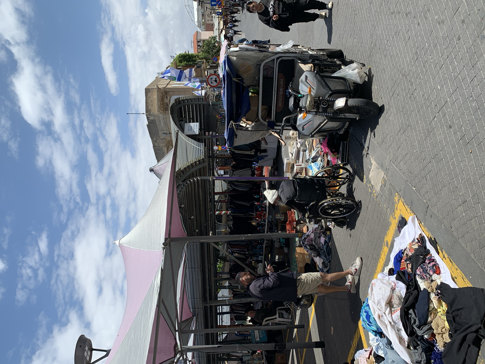 The image shows a street market in Tel Aviv, Israel. A man in a dark jacket and light shorts stands under a striped canopy, appearing to look at his phone. A person in a wheelchair is nearby, with clothing racks behind them. To the right, a pile of clothing is spread on the pavement, marked with yellow lines. A three-wheeled vehicle is parked nearby, heavily loaded with goods. Israeli flags are visible in the background, attached to buildings. The sky is blue with scattered clouds. The street is paved with interlocking blocks.