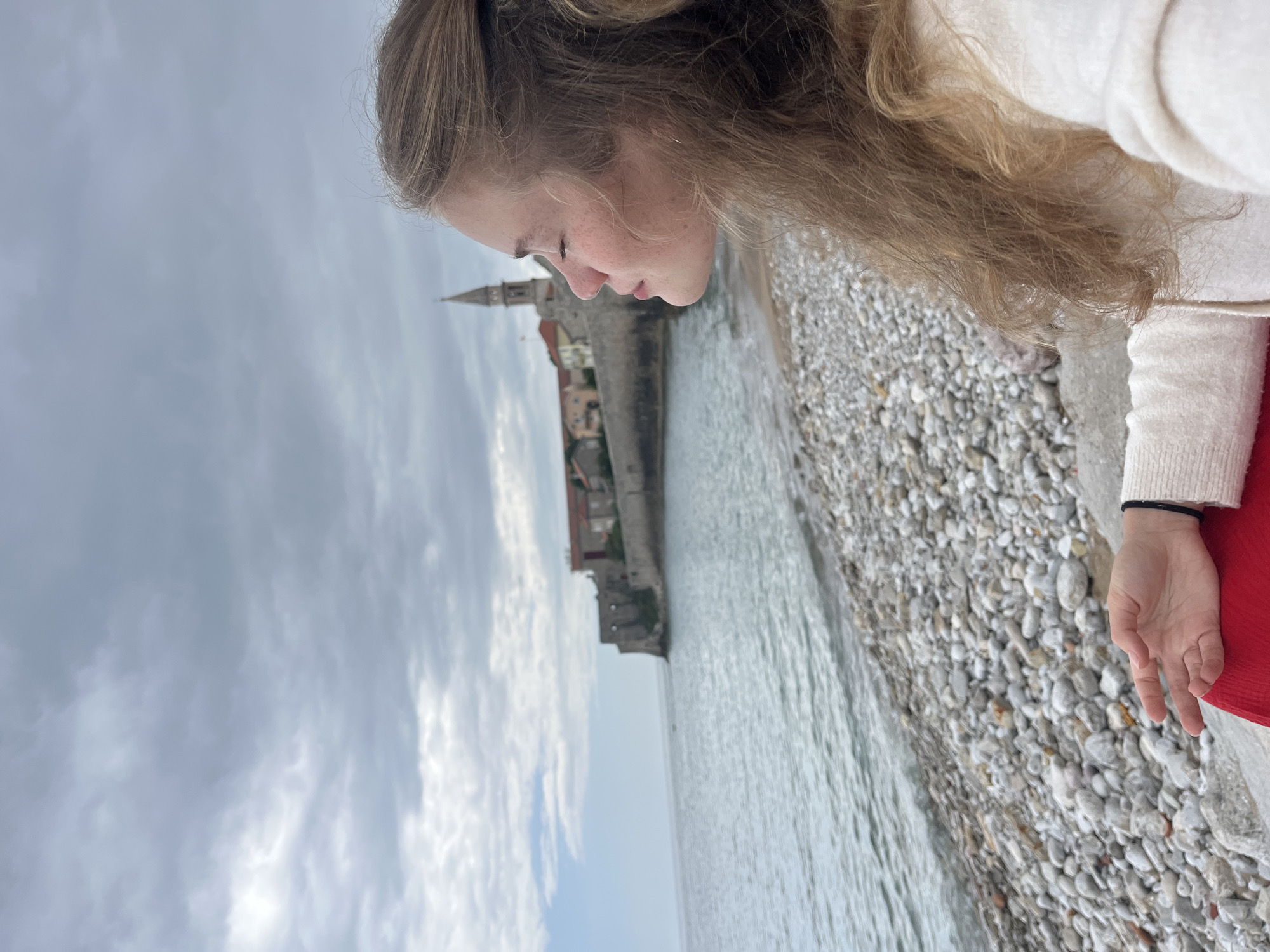 A woman stands on a pebble beach in Budva, Montenegro. She has fair skin, light brown hair pulled back, and wears glasses. She is dressed in a light-colored sweater with a red garment visible near her hand. She looks down towards the water, with the Old Town of Budva visible in the background. The sky is overcast. The beach is covered in small, rounded pebbles. The water is calm.