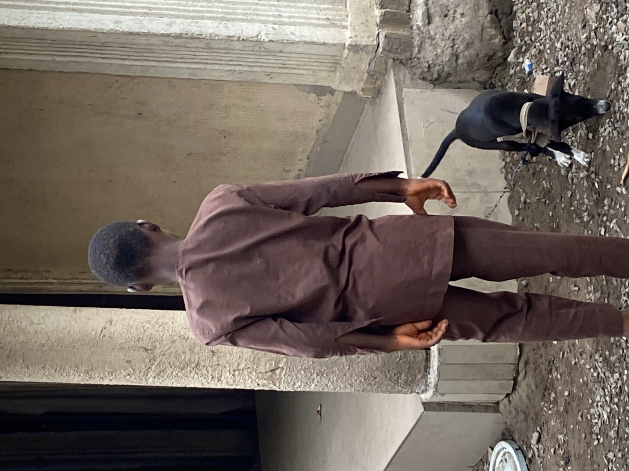 A man in a brown suit stands near a building in Mararaba, Nigeria. A black dog with a light-colored harness and leash rests on the ground nearby, in an area covered in gravel. The building's exterior is partially visible, showing a concrete structure.