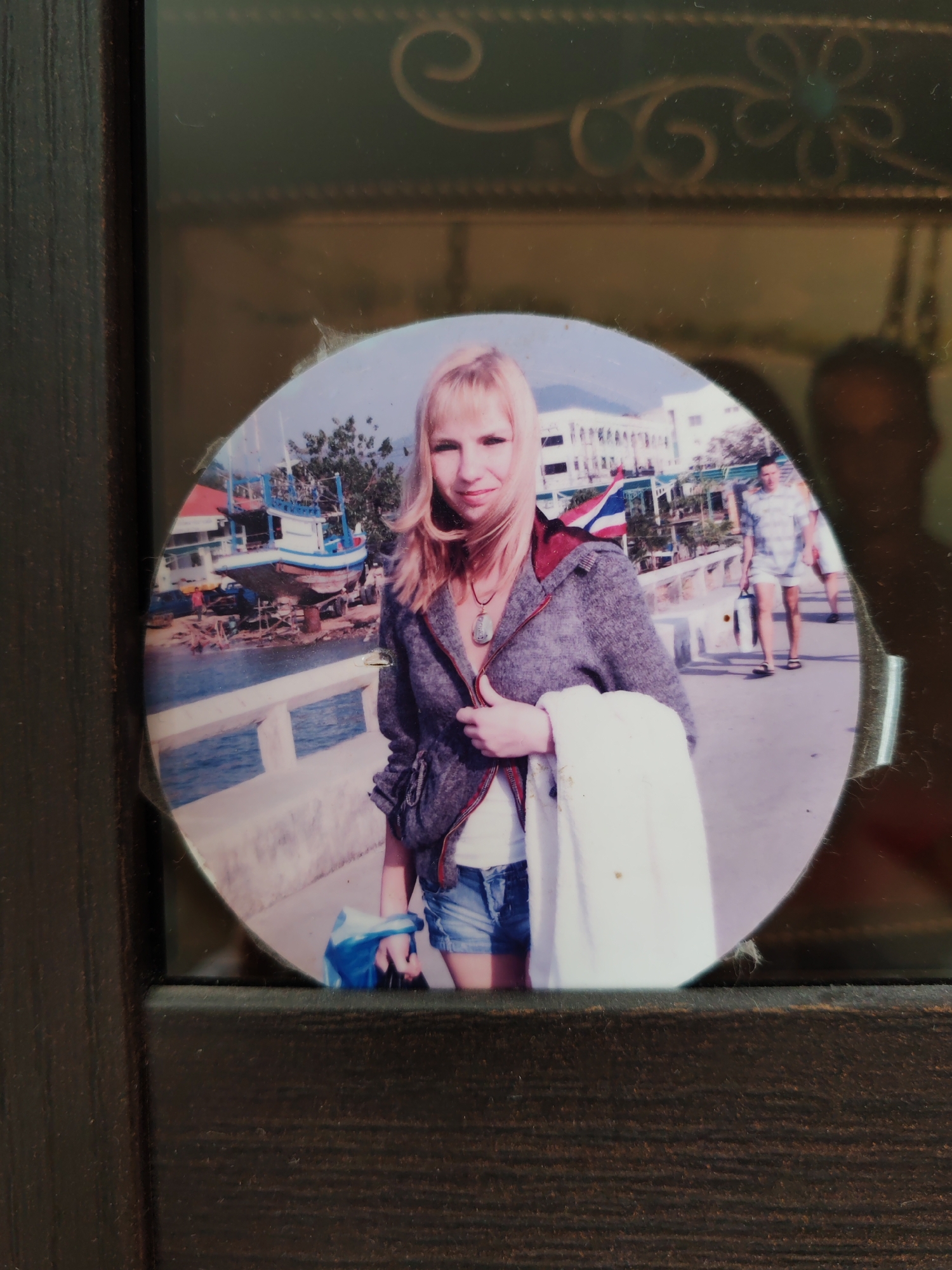 A circular physical photograph is presented within a dark brown picture frame, which features golden, ornate decorative swirls on its upper section. The main subject is a blonde woman, slightly smiling, wearing a textured grey or purple jacket over a white top and denim shorts. She has a distinctive pendant necklace and is holding a white cloth or towel over her left arm and a blue bag in her right hand.
She stands on a concrete pier or walkway beside a body of water. In the background, under bright daylight, traditional fishing boats, reminiscent of longtail boats, are visible on the shore and floating in the water, with some appearing to be under repair. Buildings and green foliage line the coastline behind the boats. Notably, a Thai flag is prominently displayed on one of the buildings in the background, strongly indicating a Southeast Asian setting, likely Thailand, which contrasts with the provided location context of Saint Petersburg, Russia.
Further to the right in the background, a man in a plaid shirt and shorts walks along the pier, carrying a white bag. Another individual is partially visible behind him. The image shows some wear, particularly around the circular edge of the photo. A faint reflection of a person is visible on the right side of the frame's glass. No legible text is visible within the photograph.
