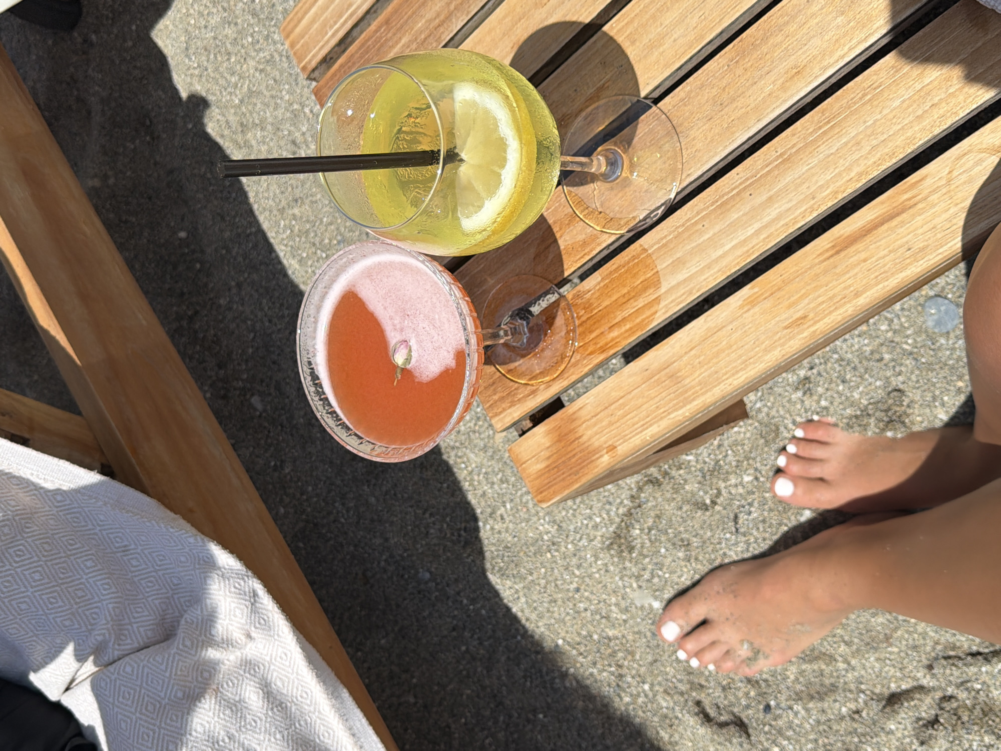 The image captures a scene at a beach in Alanya, Turkey. Two cocktails, one yellowish with a lemon slice and a black straw, and the other pinkish, sit on a small wooden table. Adjacent to the table is the sand of the beach, where a person's feet are visible. The toes have white nail polish. A part of a chair is visible along with a patterned white cloth.