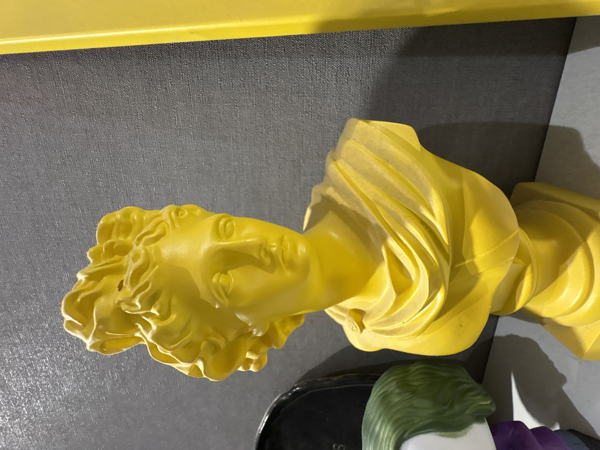 The image shows a yellow bust of a figure, possibly ancient Greek or Roman in style, with ornate hair and draped fabric around the shoulders. The bust is situated on a grey surface, with a yellow border visible at the top. In the lower right corner, there are fragmented elements of what appears to be another figure, featuring green and purple colors. The setting is a room or space, possibly indoors, though the specific location or city is unknown. There is an object on which the statue is set, maybe some sort of decorative plate or holder.