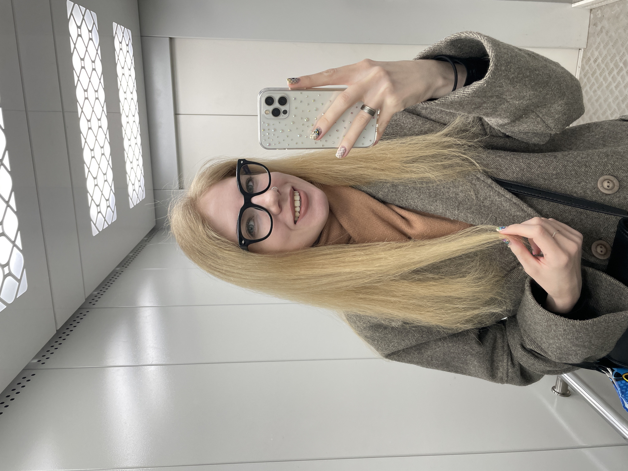 A young woman takes a selfie inside an elevator. She wears black-rimmed glasses and a brown coat. Her blonde hair is shoulder-length. She is smiling. In her hand, she holds an iPhone with a decorated case. The elevator interior is white with decorative ventilation panels. The location cannot be determined from the image.