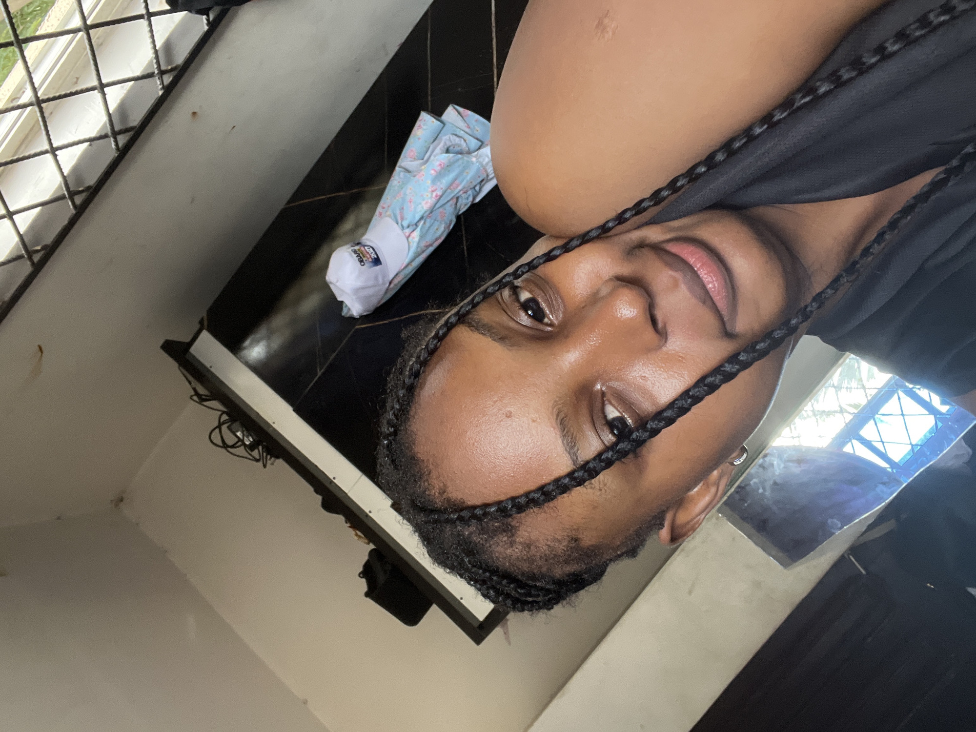 A close-up, slightly skewed, selfie of a person with braided hair and dark skin. The person is wearing a black top. The background includes a window with a metal grid, a dark-colored surface with a floral-patterned item, and a light-colored wall. The setting seems to be indoors. There is no location-specific information to confirm this photo was taken in Lokoja, Nigeria.