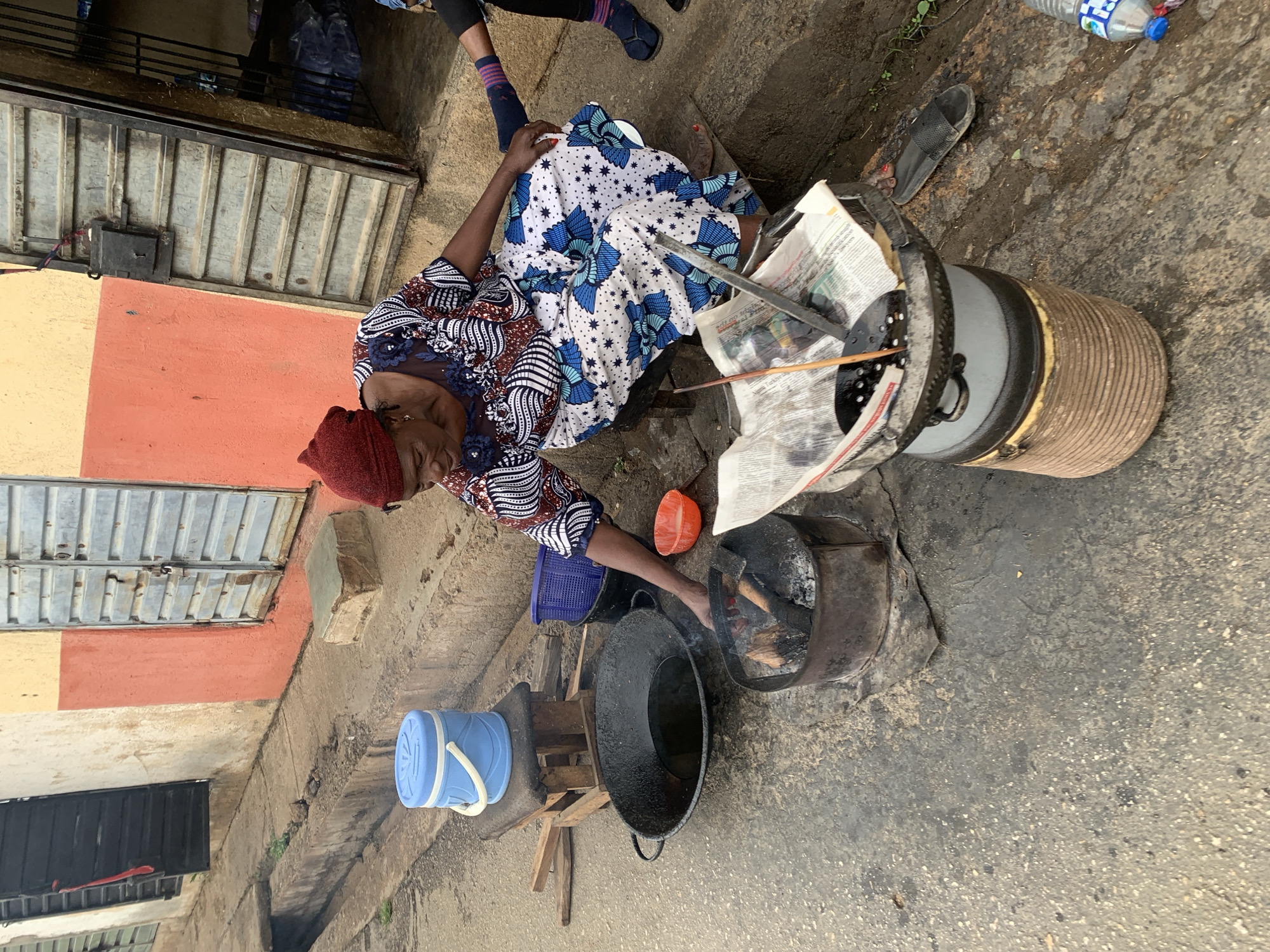 A street scene in Jos, Nigeria, shows a woman seated on the ground beside her cooking setup. She wears a red head covering, a patterned blouse with blue lace trim, and a long white skirt with blue floral designs. She faces right, attending to a cooking fire inside a blackened metal cylinder. In front of her is a large, dark wok-like pot. A white insulated cooler sits nearby on a low wooden stand. Behind her is a tall cylindrical container partially wrapped in brown ribbed material and topped with a metal lid covered in newspaper. A small orange bowl and a blue plastic strainer are also on the ground near the cooking area. The scene takes place on a paved surface beside a building with a barred metal gate and a wall painted in peach and yellow. Another person's legs and feet are visible at the top of the frame.