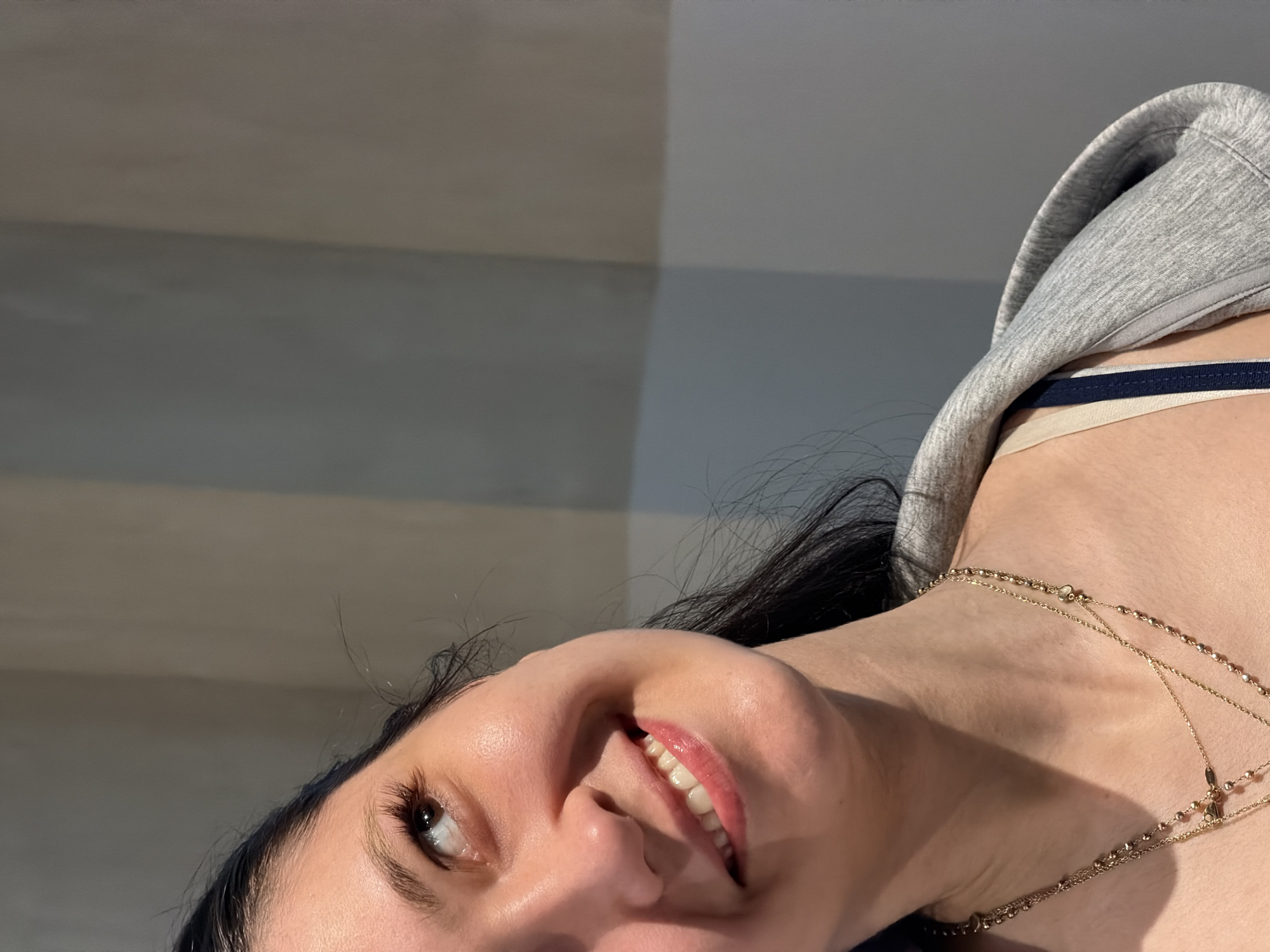 A person's face and upper torso are visible in a close-up, slightly tilted upward. The person has dark hair, fair skin, and is smiling, revealing their teeth. They are wearing a gray hooded sweatshirt, and a thin, layered gold necklace is around their neck. A bra strap with blue and white stripes is visible beneath the sweatshirt. The background is a light gray wall. The location is indeterminable.