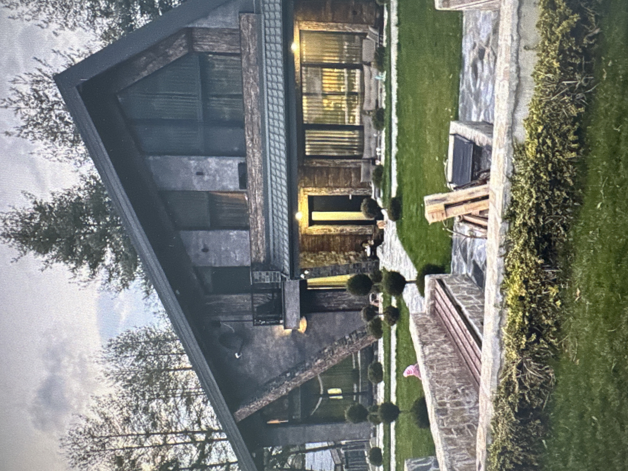 Here is a description of the image:
The image shows a modern, angular house with a dark, steeply sloped roof. The exterior is a mix of wood and what appears to be concrete or stone. Large windows are visible, and some are illuminated. The house is surrounded by a well-manicured lawn and neatly trimmed, round bushes. A stone pathway leads up to the house. A stone wall borders a section of the lawn, and a wooden bench sits adjacent to the wall. The background includes trees and a cloudy sky. The location is in a city of Unknown, Unknown. There are no people visible in the image.