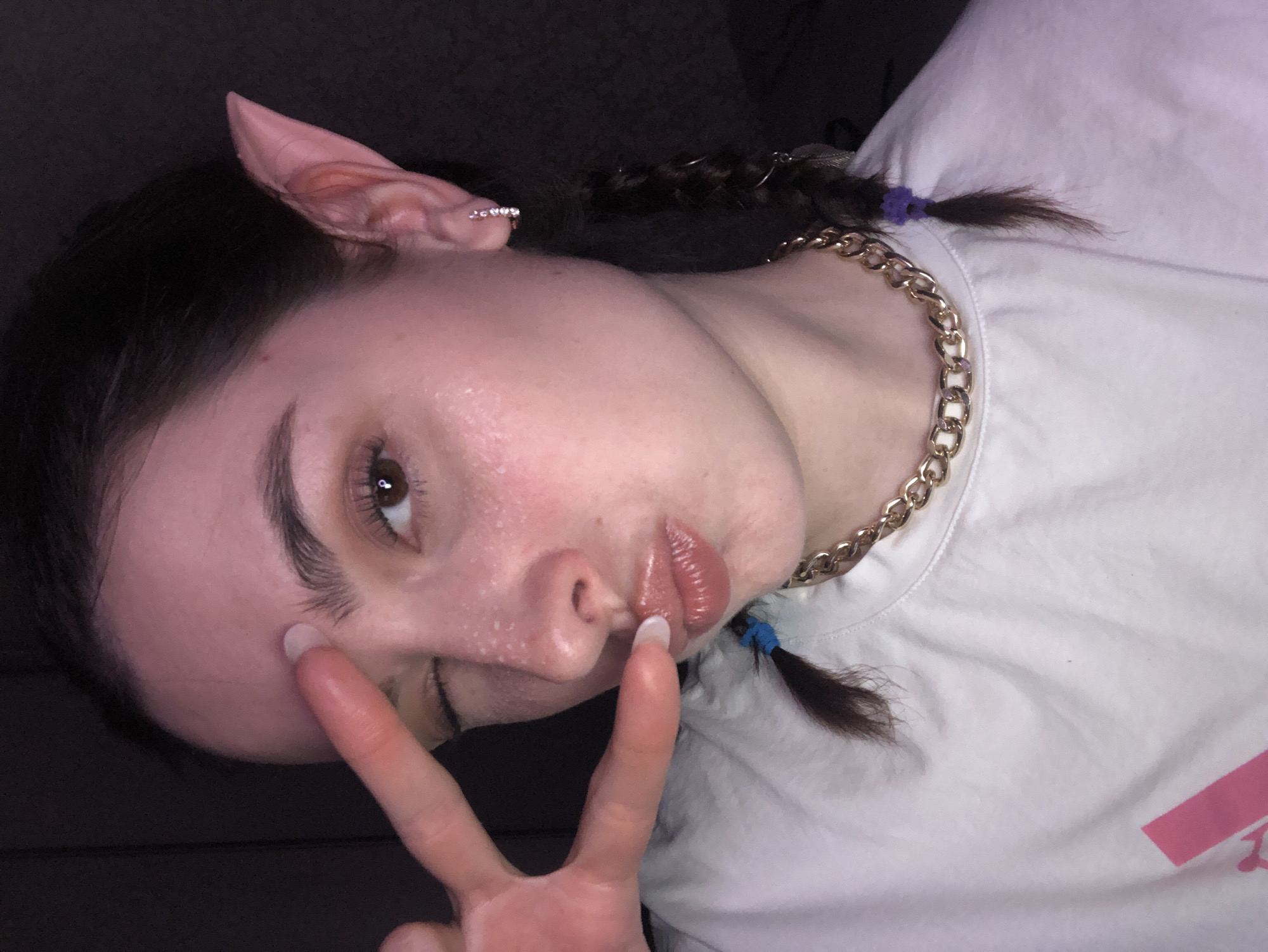 A young adult, likely female, poses for a close-up, rotated selfie indoors in Noviy, Russia. Her head is tilted to the side, and she is making a peace sign with her left hand while slightly puckering her lips. She has dark hair styled in two braided pigtails, secured with purple hair ties, and a small silver feather charm is visible in one braid. She is wearing prosthetic elf ears, one of which is clearly visible with a small, sparkly silver earring. A thick gold chain necklace is worn over a white t-shirt. There's an indistinct pink graphic or text partially visible on the bottom right of her white t-shirt. Her skin appears somewhat shiny or highlighted. The background is dark and nondescript, suggesting an indoor setting. The overall mood is playful.