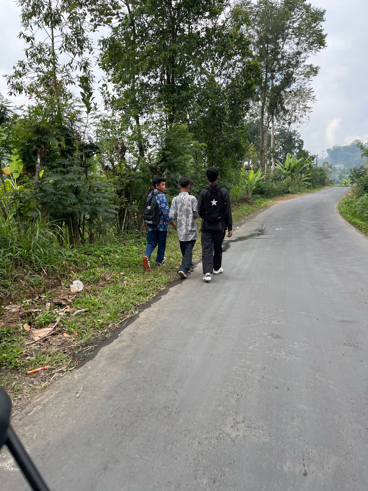 Three young males are walking away from the camera on a paved road. The road curves to the right and is flanked by a grassy shoulder on the left, lined with dense vegetation including trees and banana plants. The sky is overcast. The male on the left wears a blue backpack and a patterned shirt. The middle male wears a patterned shirt and dark pants. The male on the right wears a black jacket with a white star emblem on the back, dark pants, and white sneakers. The scene appears to be in a rural or semi-rural area.