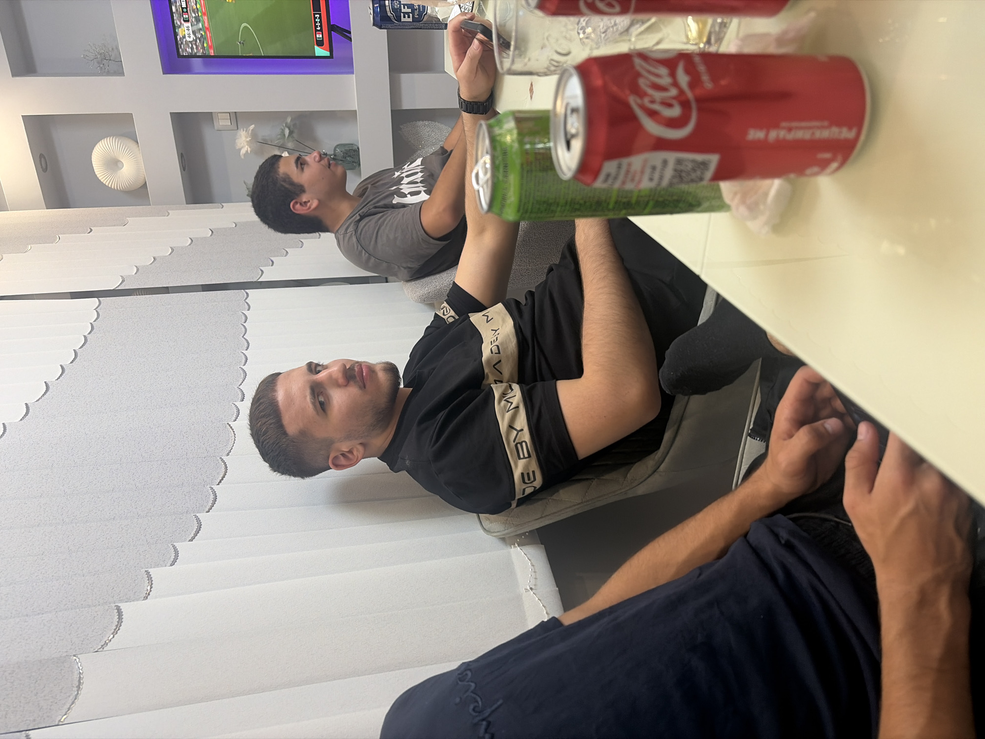 The image captures an indoor scene, likely a living room or dining area, within a residence in Podgumer, Bulgaria. The setting appears modern, with vertical blinds on the left and illuminated shelving units on the right. A television screen displaying a sports match is visible in the background.
In the foreground, three young men are seated around a light-colored table. The man closest to the viewer, on the left, is partially obscured, wearing a dark blue t-shirt. The central figure, a young man with short hair, is wearing a black t-shirt with a beige stripe and appears to be looking to his right, away from the camera. To his right, another young man in a grey t-shirt is looking towards the television.
On the table are several objects, including two open cans of soda, one red Coca-Cola can and one green can, and a glass. The presence of these items suggests a casual gathering. The overall atmosphere appears relaxed, with the individuals engaged in watching television. The lighting is moderately bright, highlighting the subjects and the immediate surroundings.