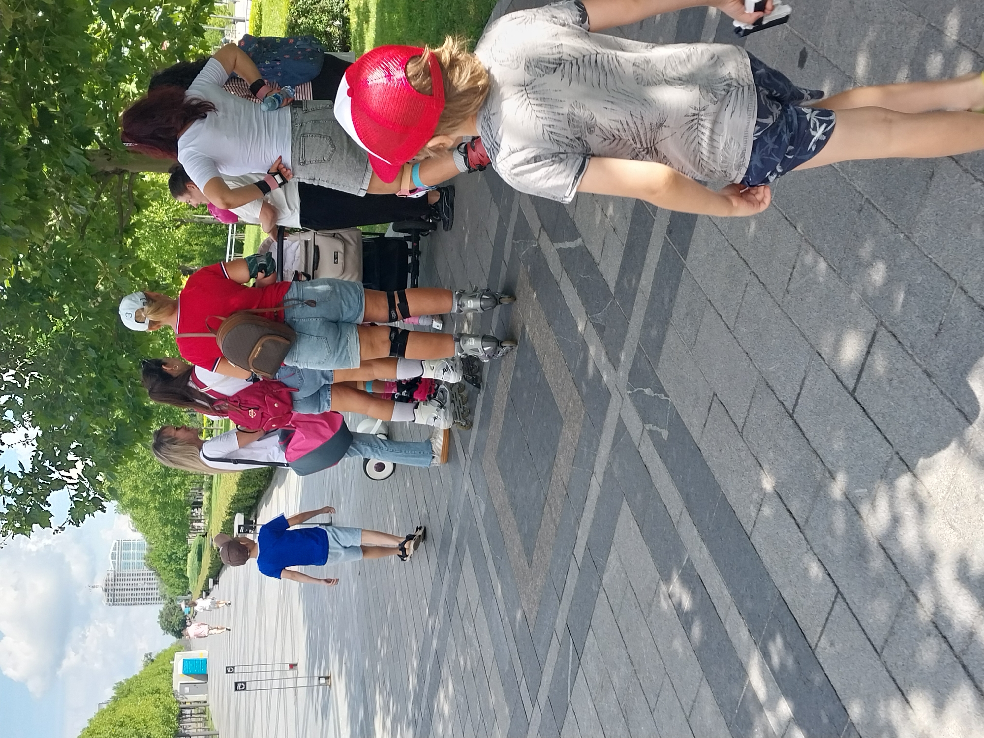 In Dnipro, Ukraine, on a paved walkway lined with trees and a green lawn, a group of people are present. The walkway has a distinctive patterned stone surface with alternating grey and darker grey sections. In the foreground, a young boy wearing a grey t-shirt with a palm leaf pattern and blue shorts with white palm prints walks away from the camera, holding a white object. Behind him, several individuals are gathered. One woman wears a red cap and a red t-shirt with denim shorts, and another woman in a white t-shirt and grey denim shorts. Others are also present in casual attire. Some individuals are wearing rollerblades. A stroller is visible amongst the group. In the background, the walkway extends into the distance, with more trees lining the sides and a tall building under construction visible. The sky is cloudy with patches of blue.
