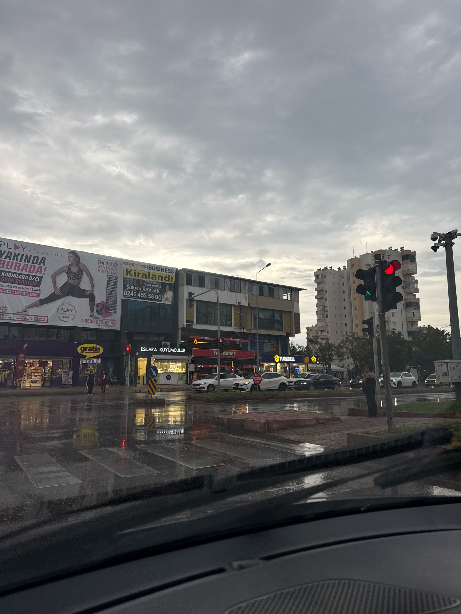 This is a photograph taken from inside a vehicle looking out at a street scene in Antalya, Turkey. The weather appears to be rainy and overcast, with wet roads reflecting the lights of surrounding businesses and buildings. A pedestrian crossing is visible on the wet asphalt, and a traffic light shows a red signal.
On the left side of the street, there are several commercial establishments. One large billboard advertises "PLAY Stüdyo Plus" with an image of a woman in athletic wear, along with text about fitness and training programs. Adjacent to this is another billboard for "REDSTONE BUSINESS Kiralandı." Below these billboards, there are storefronts, including "gratis" and "ESİLARA KUYUMCULUK" (Esilara Jewelry).
Further down the street, there are more buildings, including a multi-story apartment building. Traffic lights and streetlights are present. Several cars are on the road, some of which are white. A few pedestrians are visible on the sidewalk and crossing the street. A security camera is mounted on a pole. The overall impression is of a commercial street in a city on a cloudy and wet day.