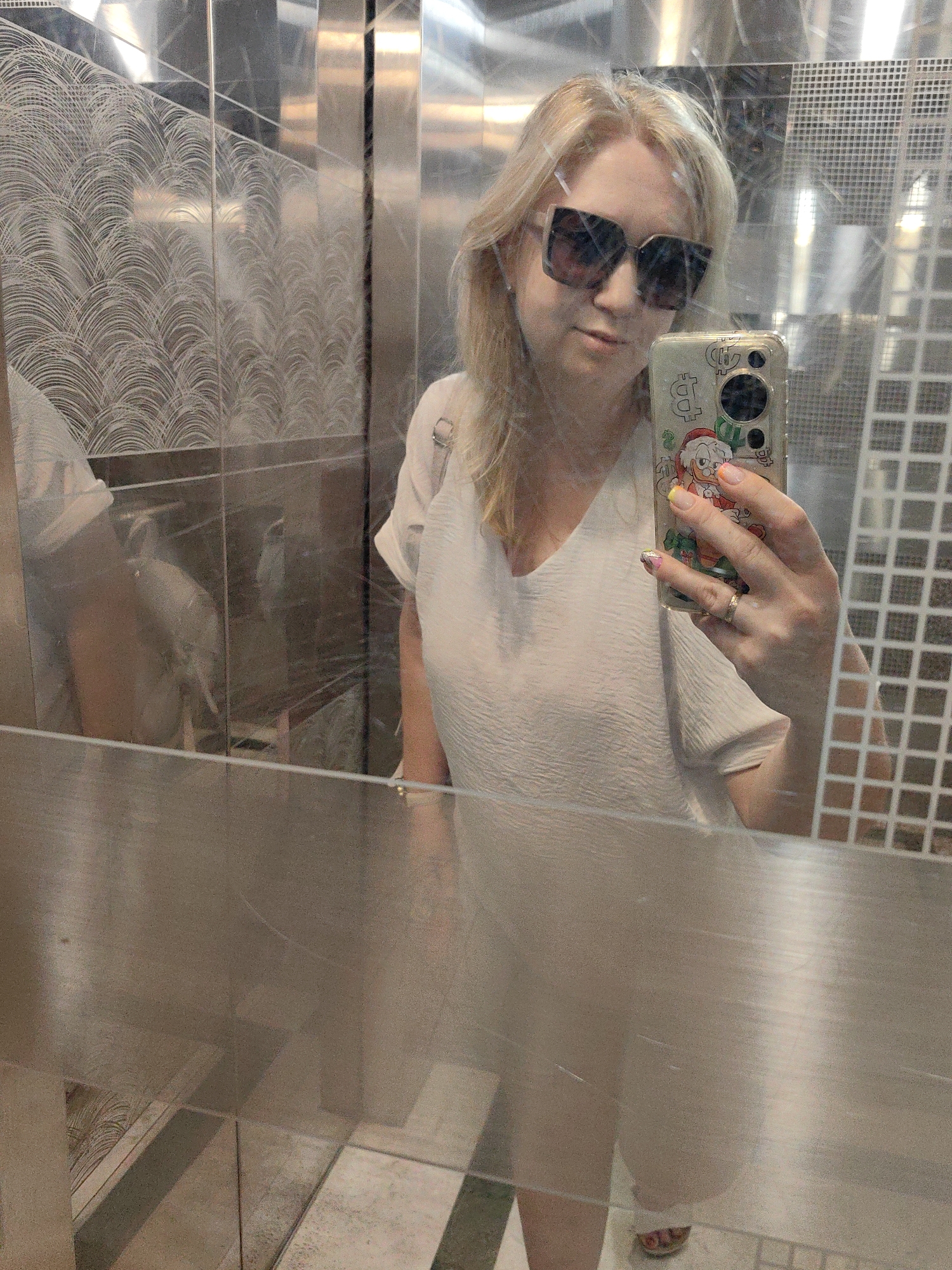 A woman with blonde hair and sunglasses takes a mirror selfie in an elevator with reflective metallic walls. She is wearing a light-colored top and holding a smartphone with a case featuring cartoon characters. Her right hand, visible in the frame, has painted nails and a ring on her finger. The elevator interior features textured metallic panels and a grid-like pattern on one wall. The lighting suggests an indoor setting. The city of Sochi, Russia is not visible in the image.