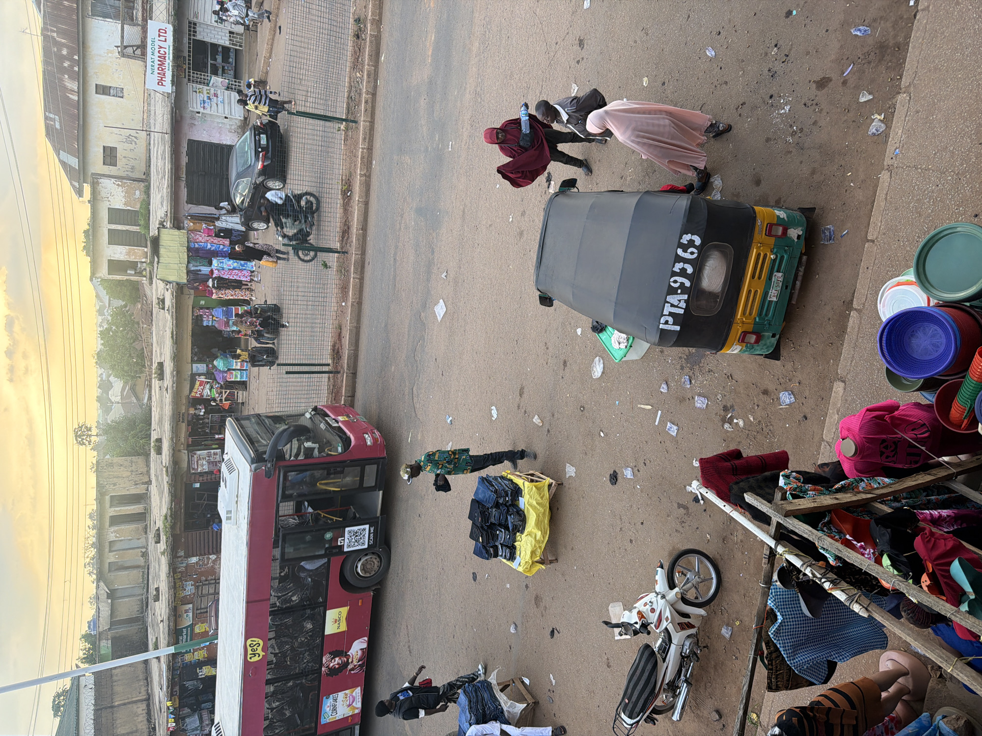 This image depicts a bustling street scene in Bukuru, Nigeria, likely during the late afternoon or early evening, as suggested by the warm, golden light illuminating the sky and casting long shadows. The scene is filled with a variety of subjects and activities. In the foreground, an autotricycle, also known as a "keke," with the license plate PTA-9363, is parked on the dusty road. Nearby, a large red bus, possibly a public transport vehicle, is stopped, its side adorned with advertisements including "Yes!" and "CORNFLAKES."
Several people are visible engaged in various activities. Two individuals are seen interacting near the autotricycle, one appearing to be wearing a dark outfit and the other a lighter-colored garment. Further down the street, a person in a green and black patterned shirt and a white cap stands next to a stall piled high with folded blue items, possibly jeans. Another person, dressed in a dark suit and cap, is crouched beside a display of folded clothes. On the right side of the image, a makeshift stall displays an assortment of colorful textiles and clothing items, with several large plastic containers stacked behind it.
The setting appears to be a commercial area, with buildings lining the street. One building prominently features a sign for "NERAT MODEL PHARMACY LTD." The street is unpaved and littered with discarded papers and debris, indicative of a busy urban environment. The overall impression is one of everyday life unfolding in a lively Nigerian town, with commerce, transportation, and pedestrian activity all contributing to the vibrant atmosphere.
