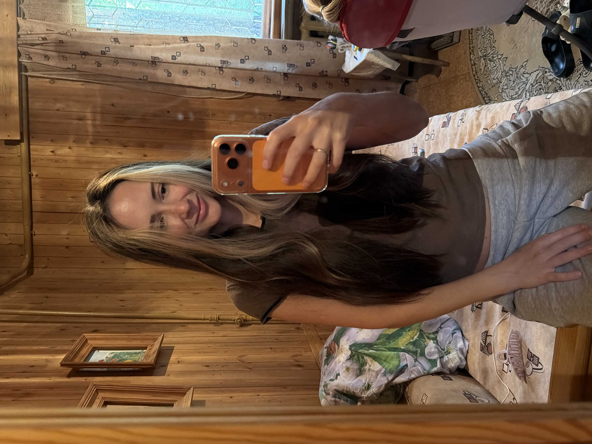 A woman with long, blonde-streaked brown hair takes a selfie in a room with wood-paneled walls. She is wearing a brown t-shirt and grey sweatpants. She holds an orange-cased smartphone in her right hand, and a ring is visible on her left ring finger. Behind her, a floral-patterned blanket is draped over a sofa. To the right, a window with patterned curtains is visible, and a radiator can be seen below it. A small table with various items, including pens and a mug, is also in the background. Black flip-flops are on the floor. The scene appears to be in a residential setting, likely a home in Udelnaya, Russia, given the context.